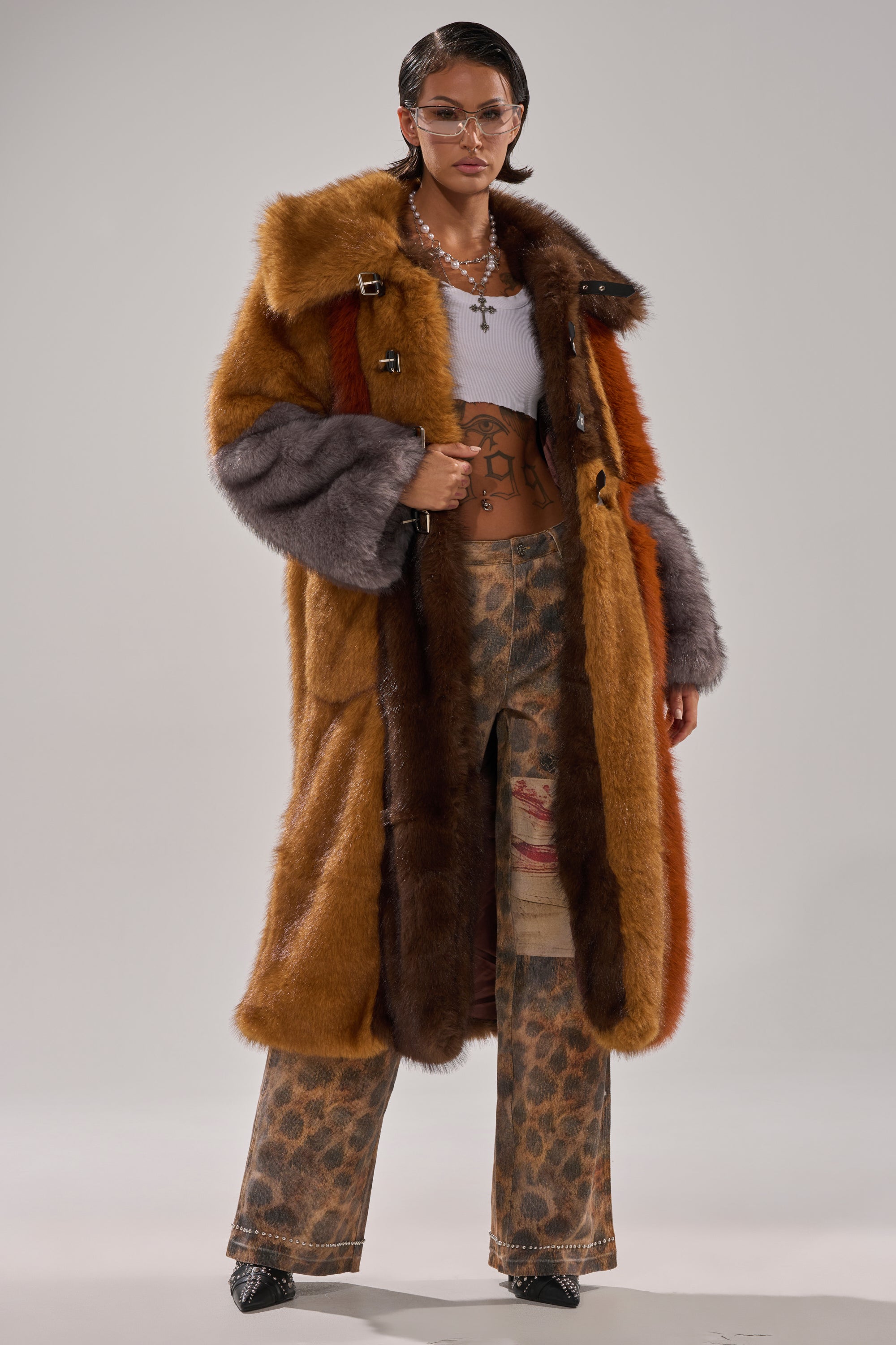 Wearing the WILD CARD PATCHWORK FAUX FUR coat over a white crop top and patterned pants, accessorized with sunglasses, layered necklaces, and earrings, a person poses confidently against a plain background.