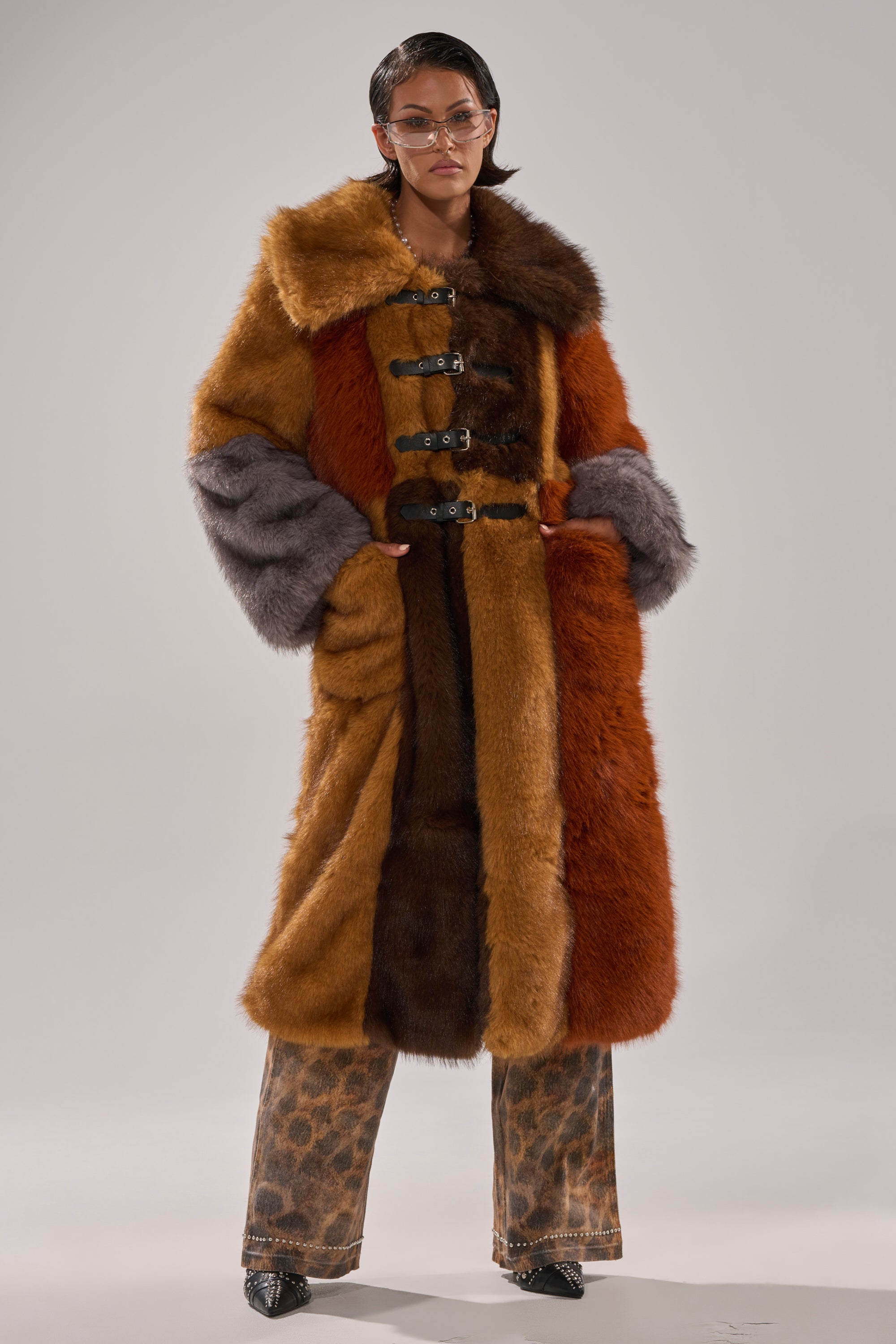 A person stands against a plain light gray background wearing the WILD CARD PATCHWORK FAUX FUR coat with large lapels and toggle closures, styled with leopard print pants and black pointed shoes.