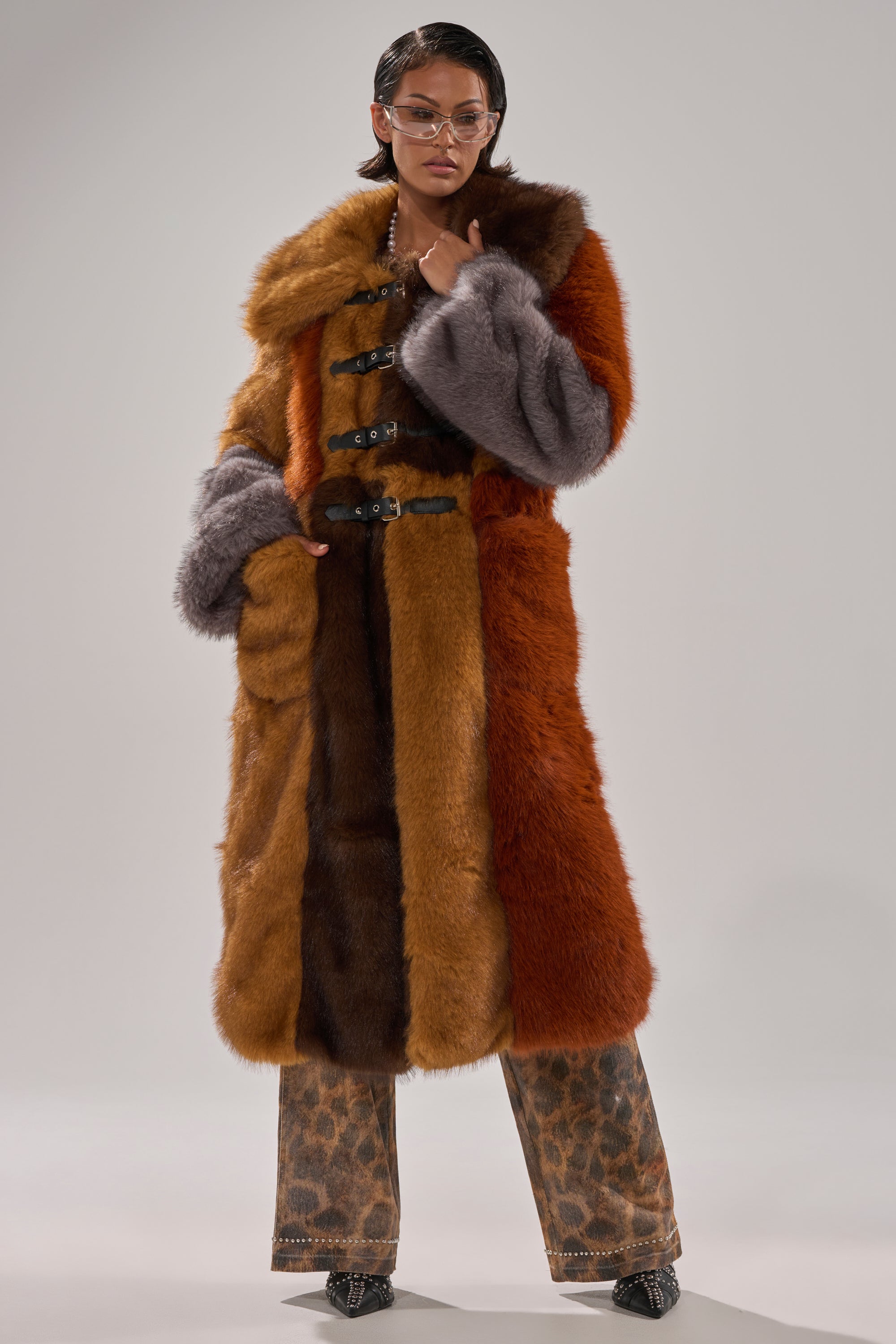 A person wears the WILD CARD PATCHWORK FAUX FUR coat featuring orange, brown, and gray panels with leopard print pants, glasses, and pointed shoes, posing confidently against a plain background.