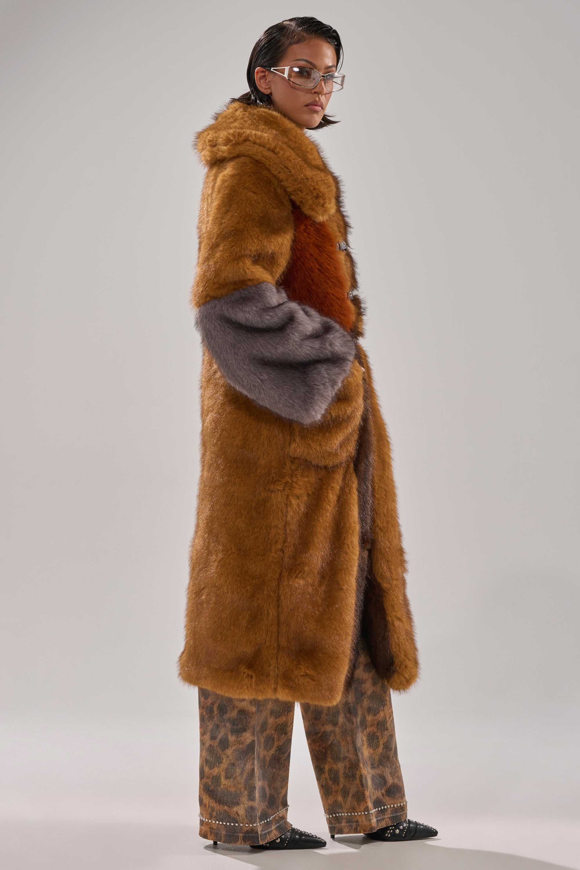 A person stands sideways with hands in pockets against a plain background, wearing glasses, patterned pants, pointed shoes, and the WILD CARD PATCHWORK FAUX FUR statement coat in long brown and gray faux fur.