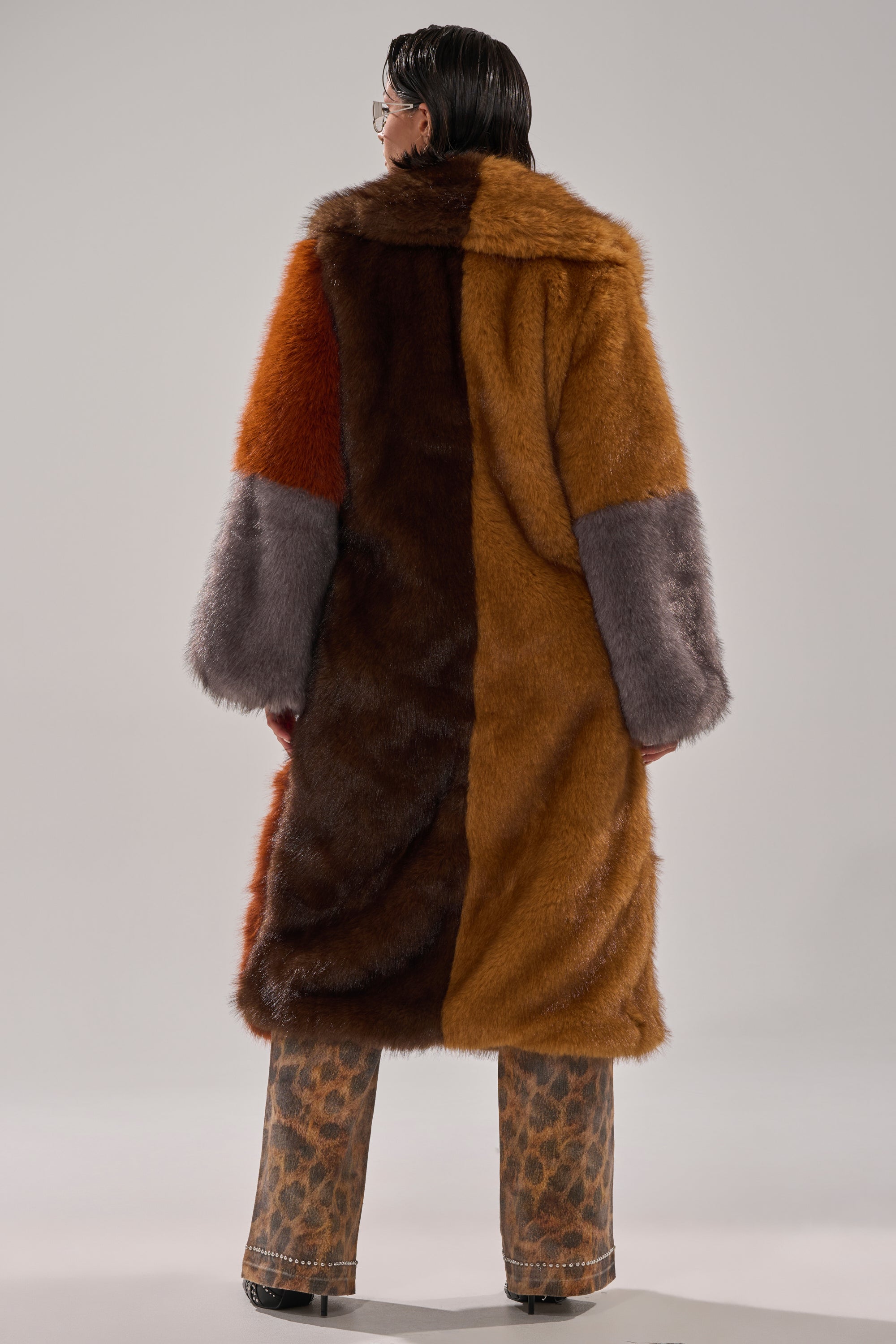 A person faces away, wearing the WILD CARD PATCHWORK FAUX FUR coat in brown, orange, and gray tones with leopard print flared pants and heeled boots. The background is plain and light-colored.