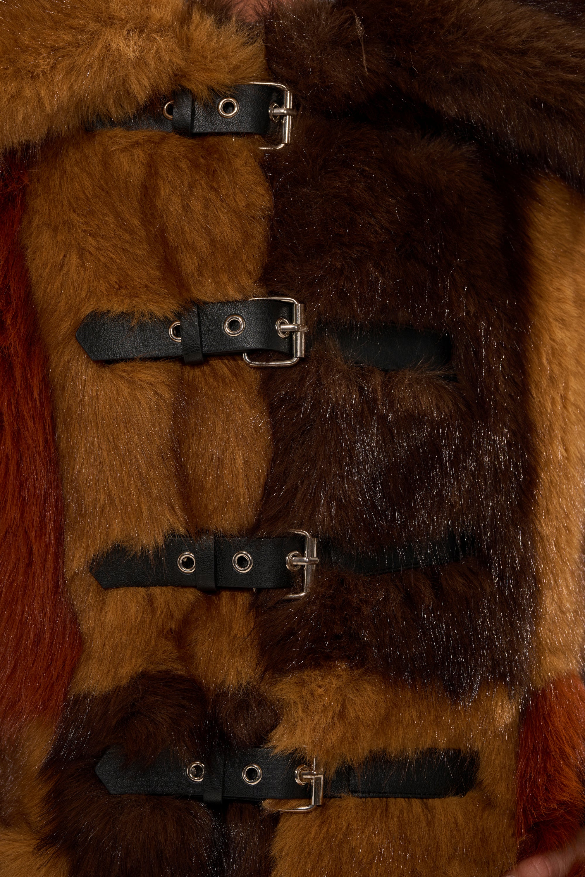 A close-up of the WILD CARD PATCHWORK FAUX FUR coat, showcasing its brown and reddish striped pattern, secured with four black leather straps and silver buckles.