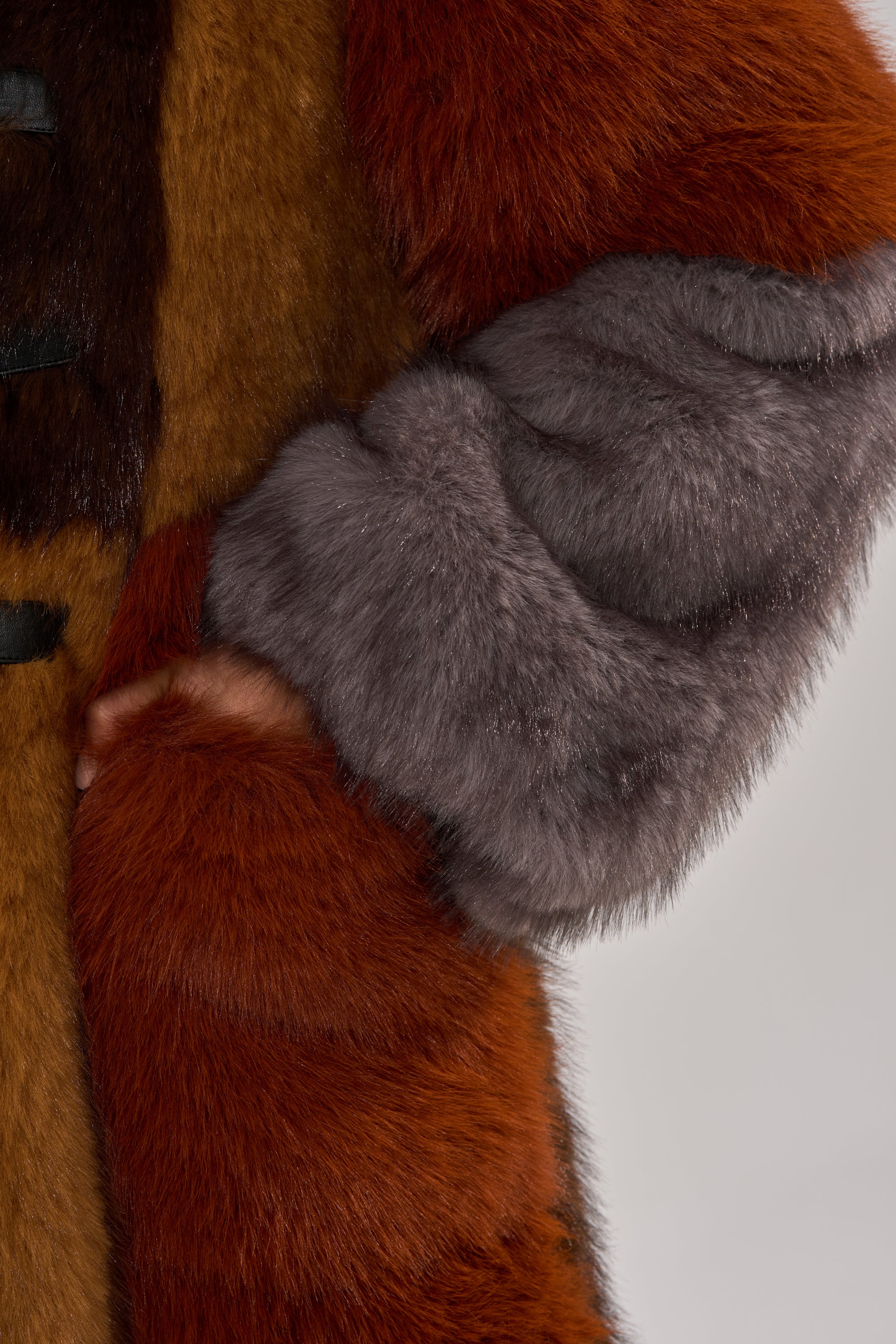 A close-up of a person wearing the WILD CARD PATCHWORK FAUX FUR coat, featuring reddish-brown and gray faux fur and bold textures. Their hand is tucked into a pocket, showcasing the coat's luxurious statement style.