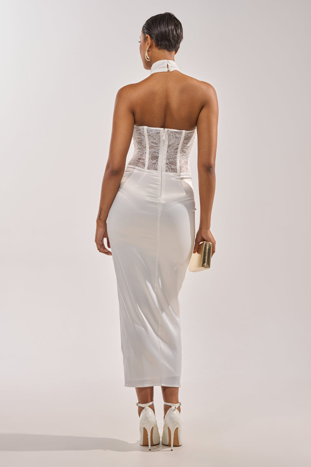 A woman faces away, wearing the EVANGELINA SATIN MAXI DRESS IN WHITE for a chic bridal look. She holds a gold clutch and wears white ankle-strap heels. Her hair is short and sleek against a plain background. 5