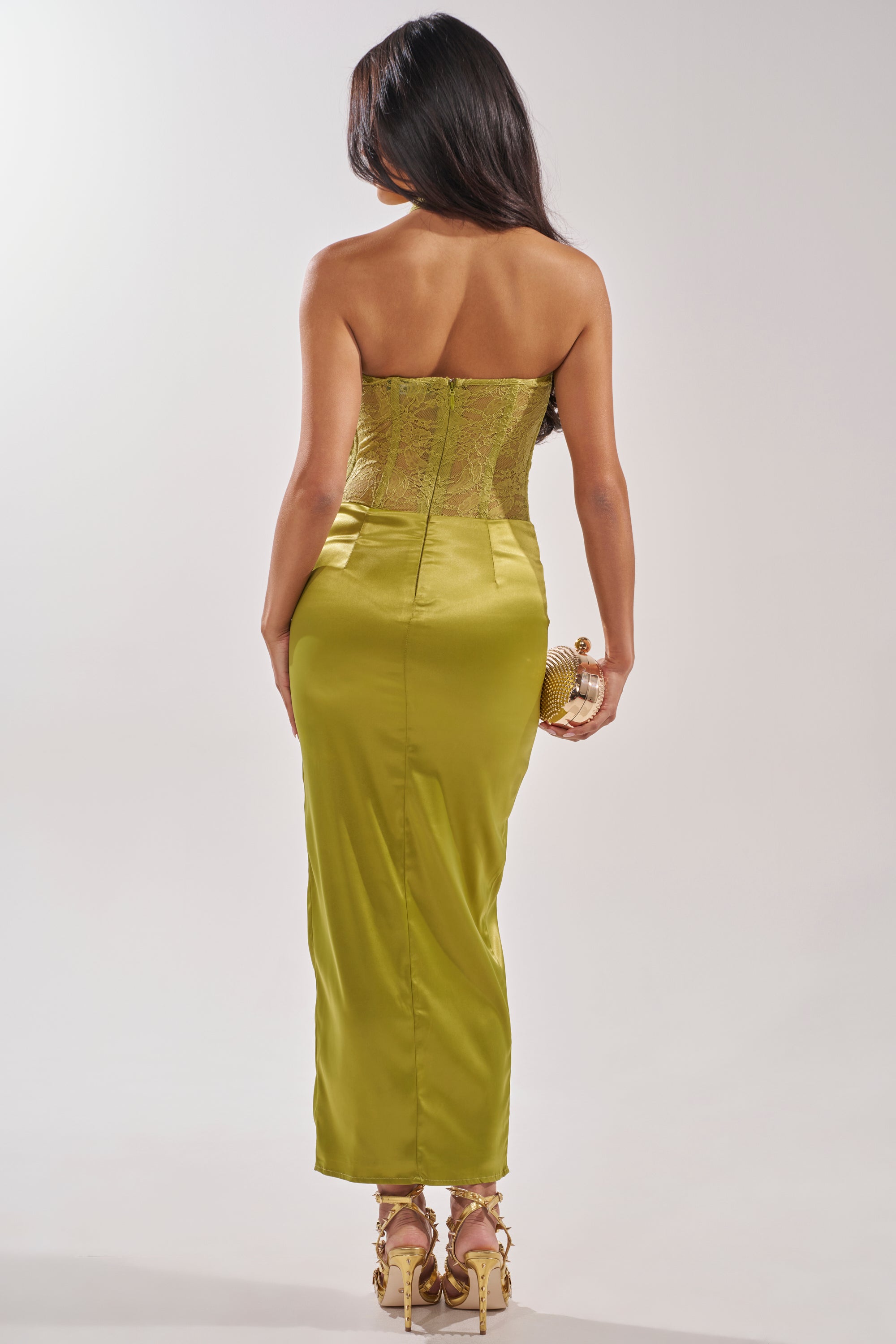 A woman with long dark hair faces away, wearing the green EVANGELINA SATIN MAXI DRESS. The strapless lace bodice and satin skirt are paired with a gold clutch and gold heels, set against a plain white background. 4