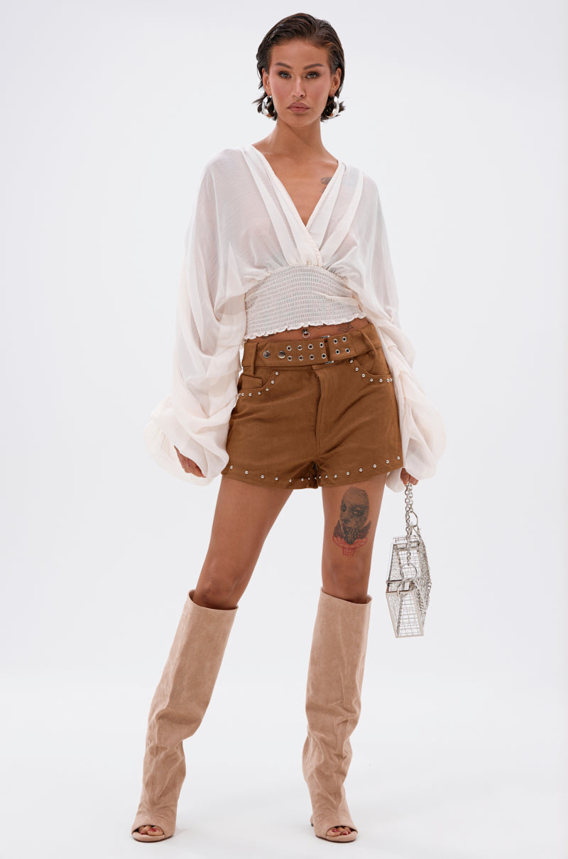 A woman stands confidently in a loose white blouse, FIGURE IT OUT FAUX SUEDE SHORTS, beige knee-high boots, and holds a small metallic handbag. She displays a tattoo on her left thigh against a plain white background. 0