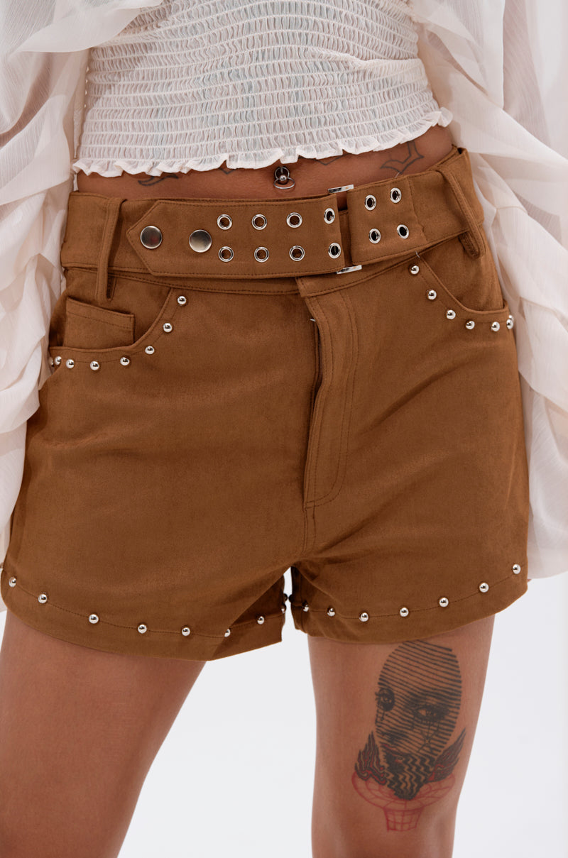 Wearing FIGURE IT OUT FAUX SUEDE SHORT in high-rise brown with silver studs, a matching studded belt, and a white textured top. A thigh tattoo of a stylized face and lines completes the look. 1