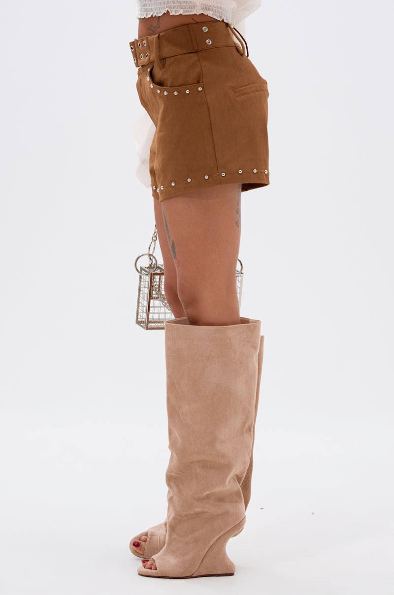 A person stands in profile, wearing high-rise FIGURE IT OUT FAUX SUEDE SHORTS with silver studs, tan suede slouchy knee-high boots, and holding a small silver cage purse against a plain white background. 4