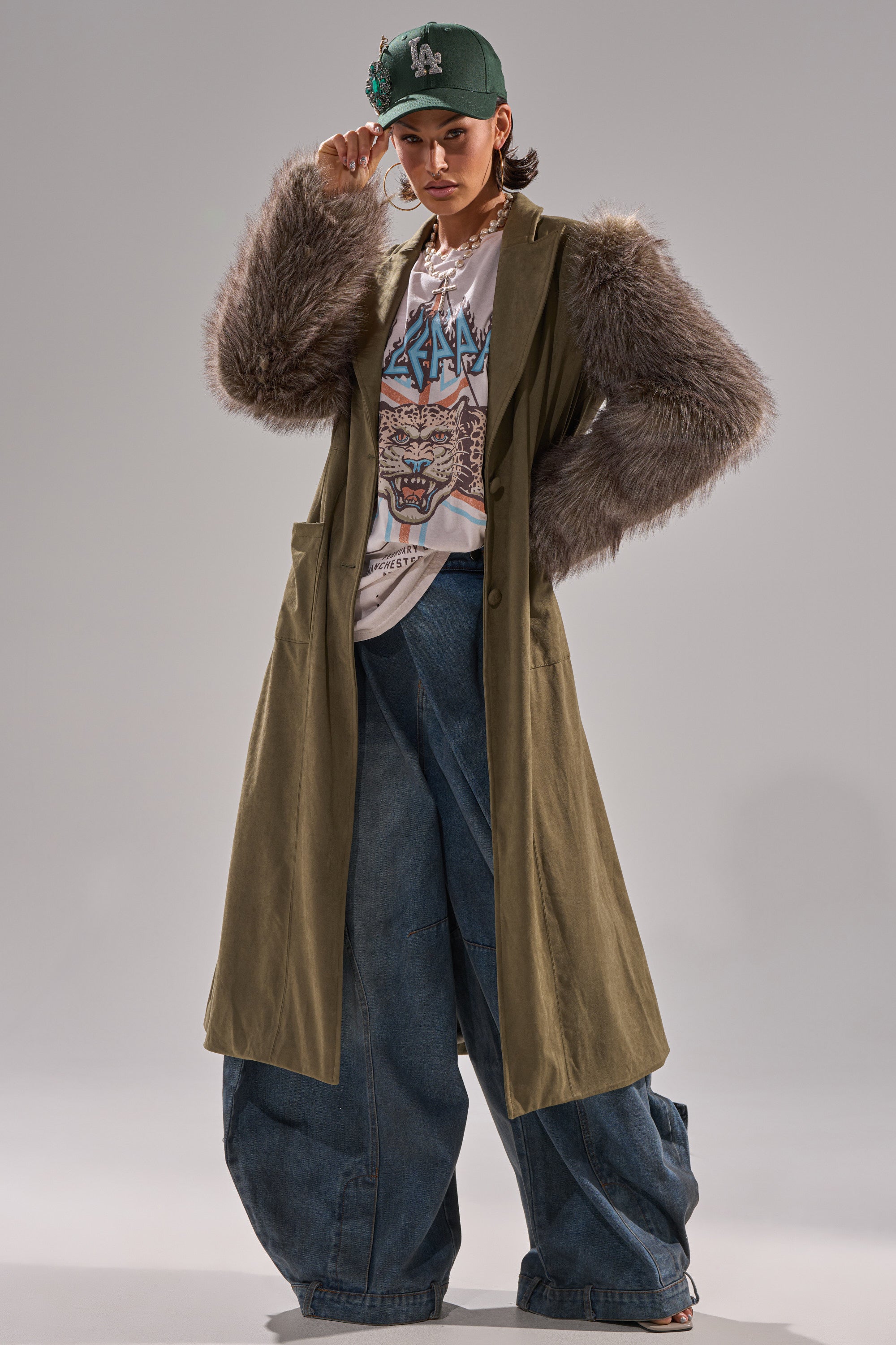 A person wearing a green cap, graphic T-shirt, the LOLITA FUR SLEEVE TRENCH, baggy denim jeans, and hoop earrings poses confidently against a plain background.