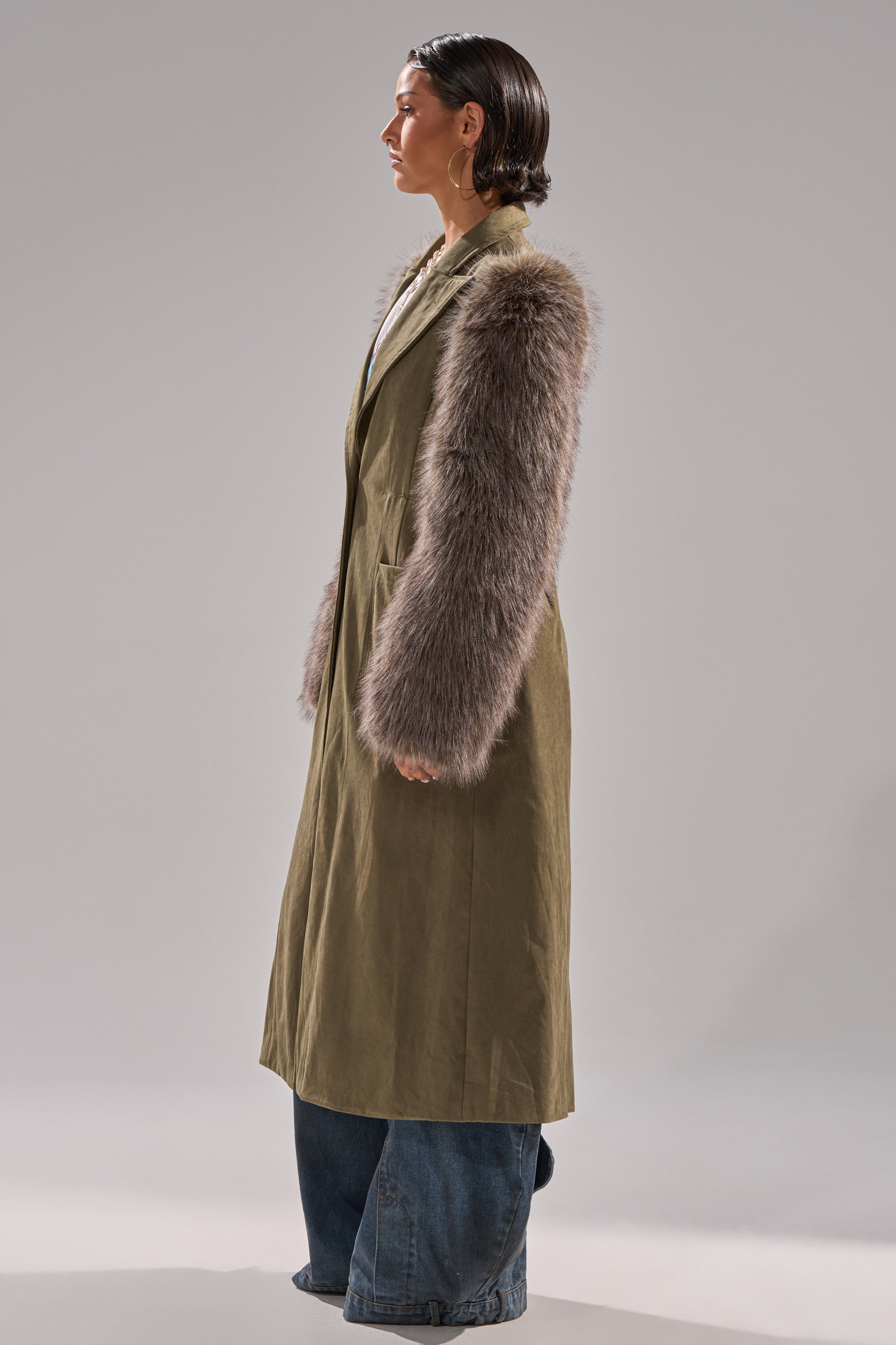 A person in profile wears the LOLITA FUR SLEEVE TRENCH—an olive green faux suede coat with shaggy faux fur sleeves—paired with wide-leg blue jeans against a plain gray background.