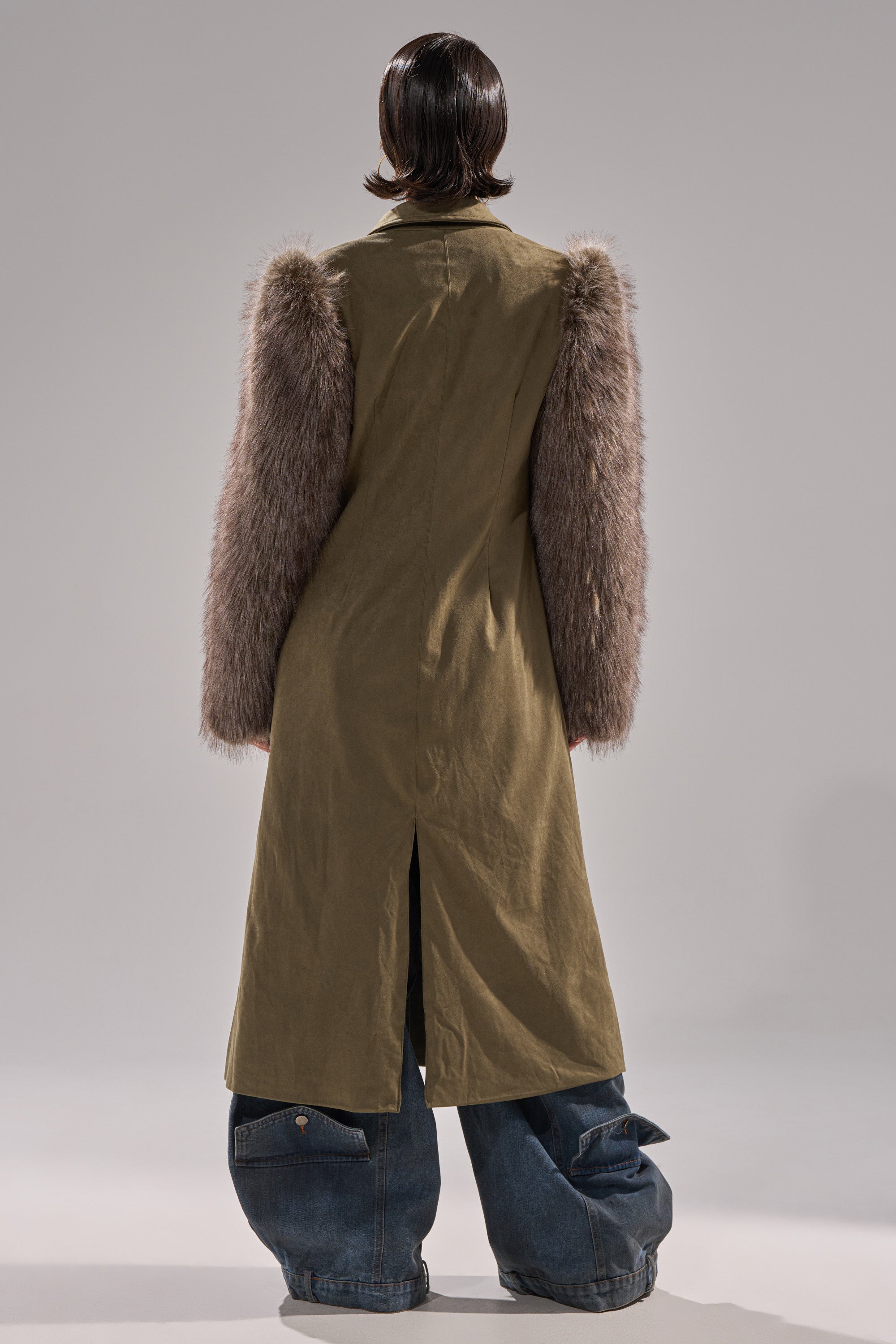 A person faces away, wearing the LOLITA FUR SLEEVE TRENCH in olive green faux suede with faux fur sleeves, paired with loose, wide-leg blue jeans against a plain neutral background.