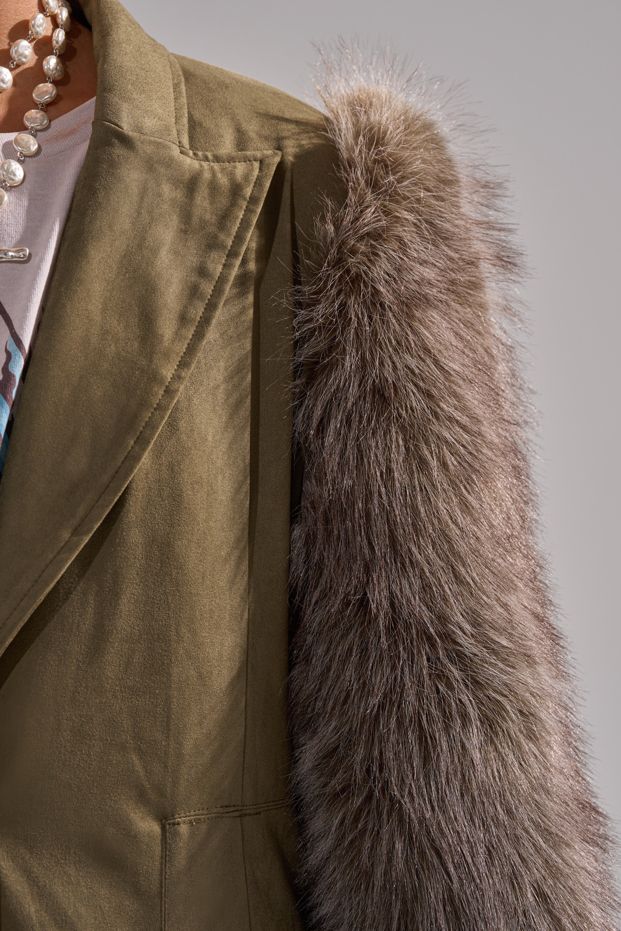 Close-up of a person in the LOLITA FUR SLEEVE TRENCH with khaki fabric and gray faux fur sleeves, layered over a white graphic tee and styled with a pearl necklace. The background is plain and neutral.