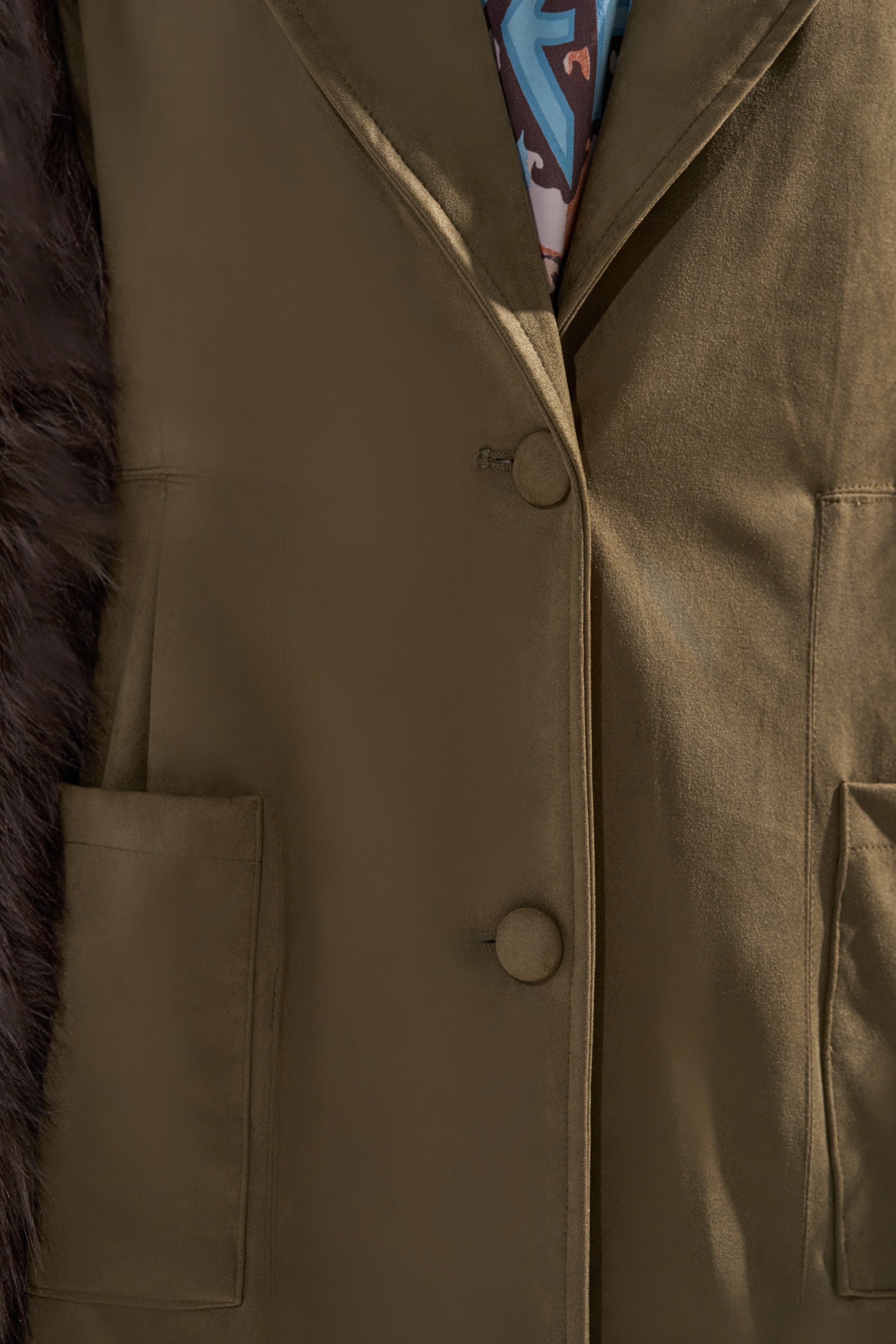 Close-up of an olive green blazer with two buttons and large front pockets, partially revealing a patterned shirt. A section of dark brown faux fur sleeve from the LOLITA FUR SLEEVE TRENCH appears on the left side.