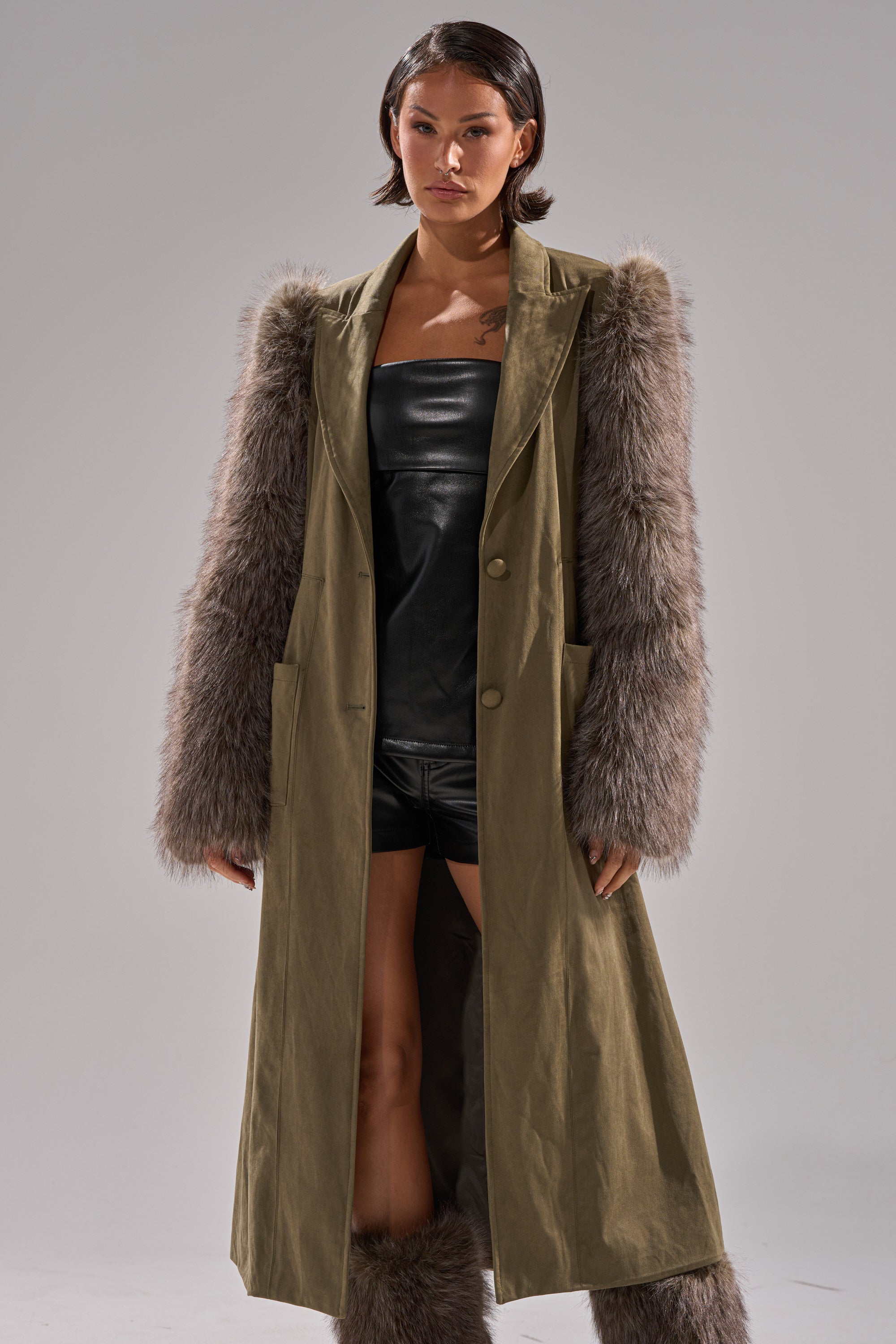 A woman with short dark hair models the LOLITA FUR SLEEVE TRENCH, showcasing its large gray faux fur sleeves over a black leather outfit and matching fur boots, posed against a plain gray background.