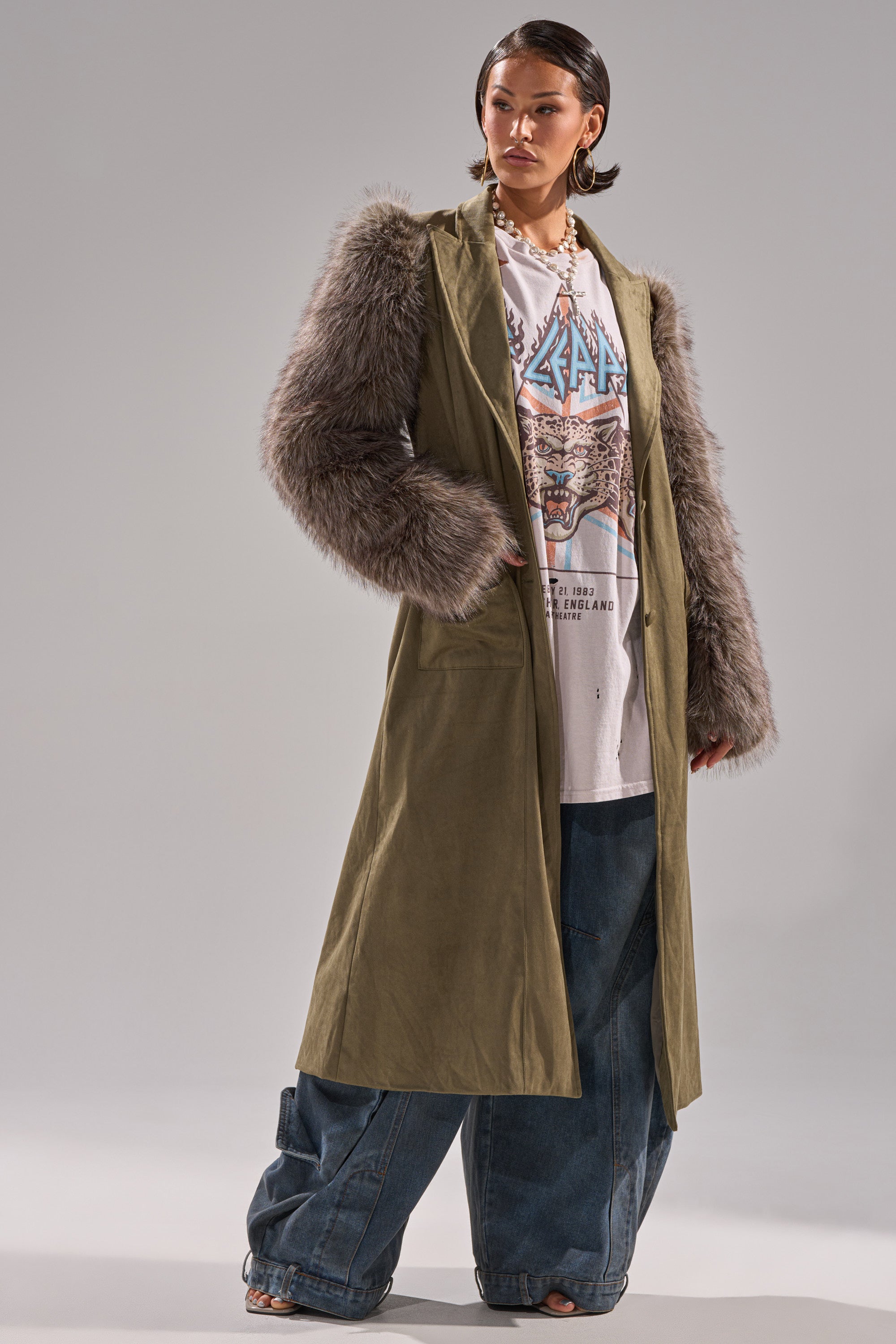 A woman models the LOLITA FUR SLEEVE TRENCH in olive green with faux fur sleeves, paired with a white graphic tee and wide-leg blue jeans, standing barefoot against a plain light gray background.