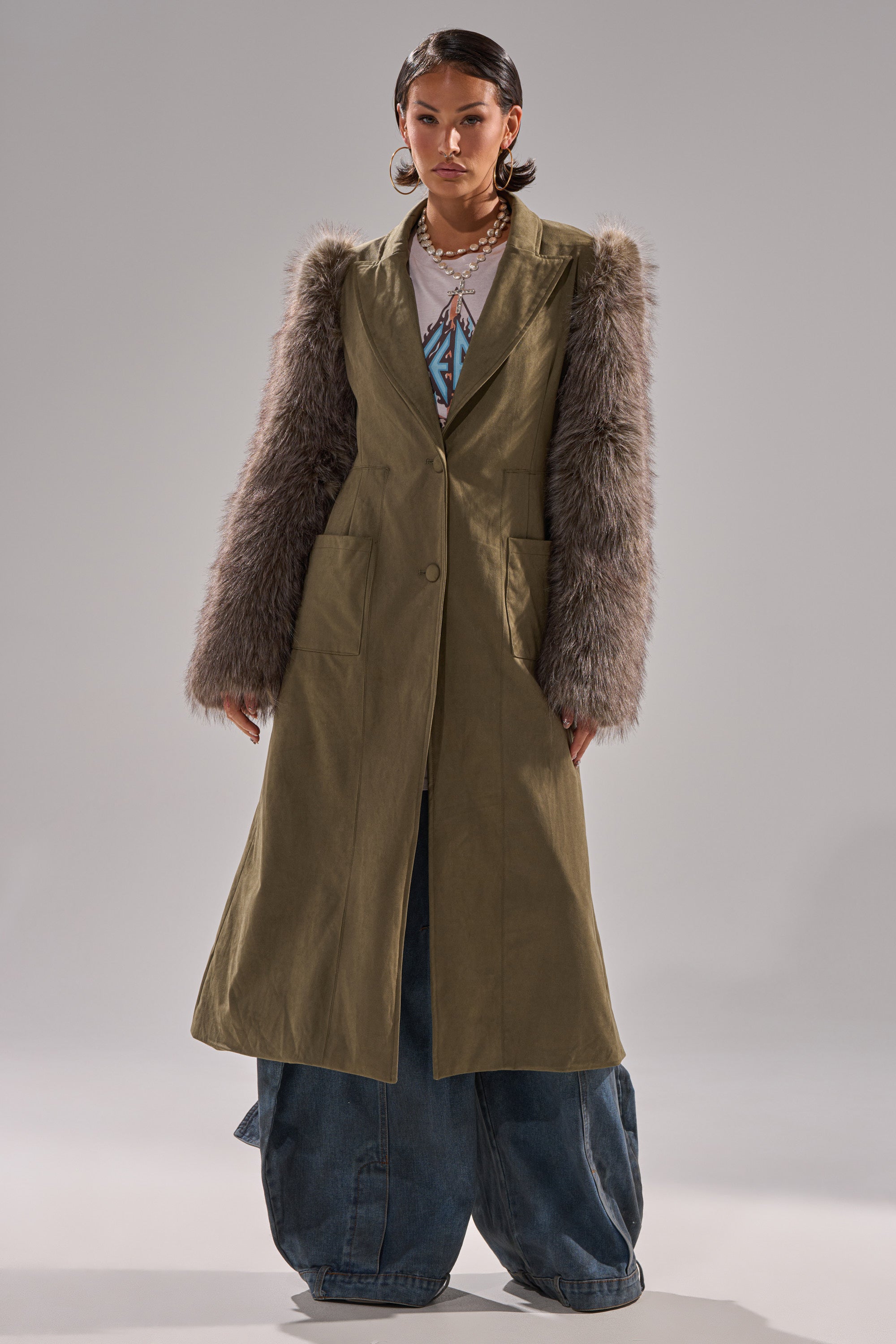 A person faces forward in the LOLITA FUR SLEEVE TRENCH, featuring shaggy gray faux fur sleeves, paired with oversized blue jeans, layered necklaces, and a white top with a blue geometric pattern against a plain light gray background.