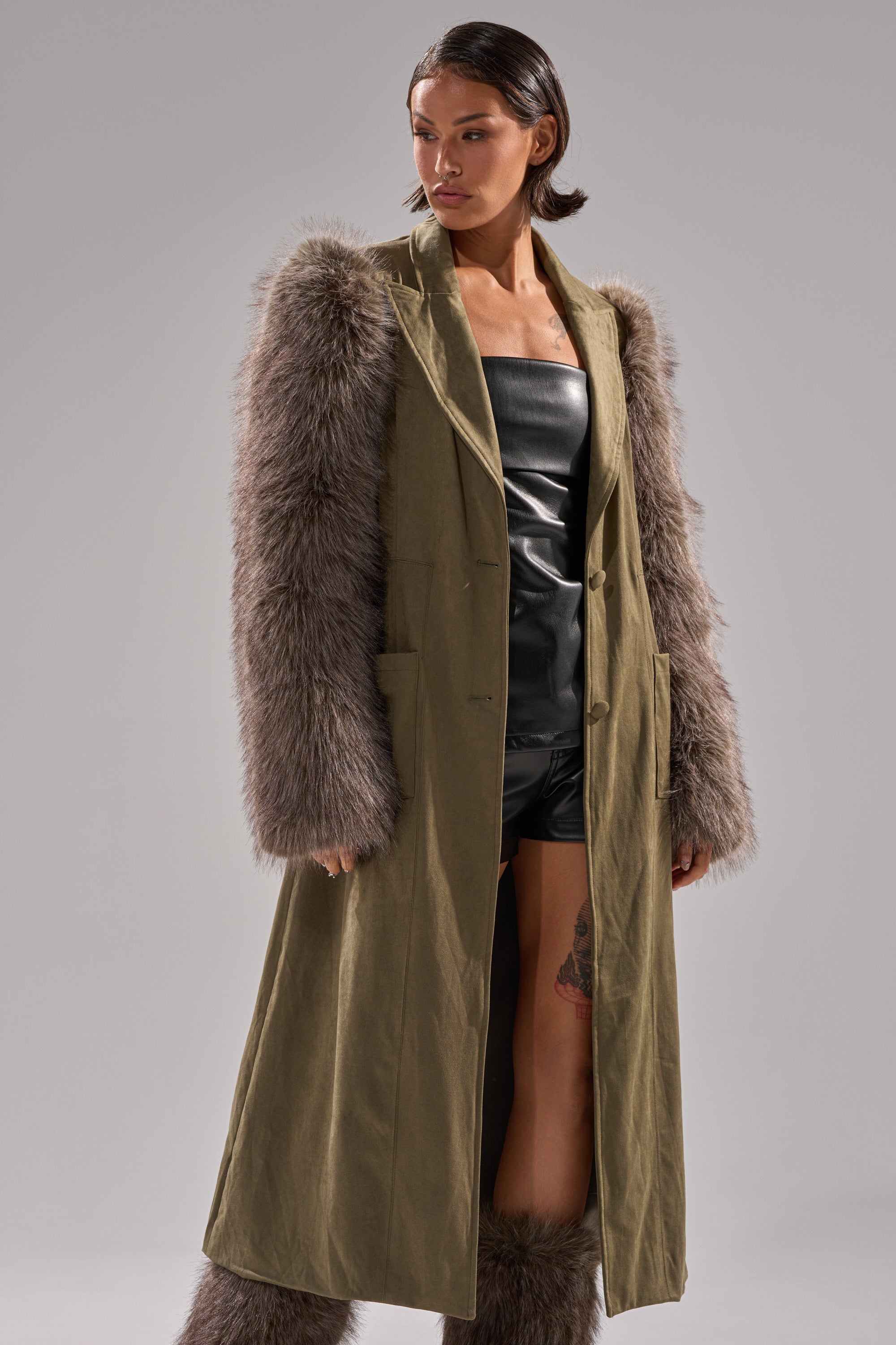 A woman stands confidently against a plain gray background, wearing the LOLITA FUR SLEEVE TRENCH—a midi-length trench coat with shaggy fur sleeves—over a black leather strapless top and shorts.