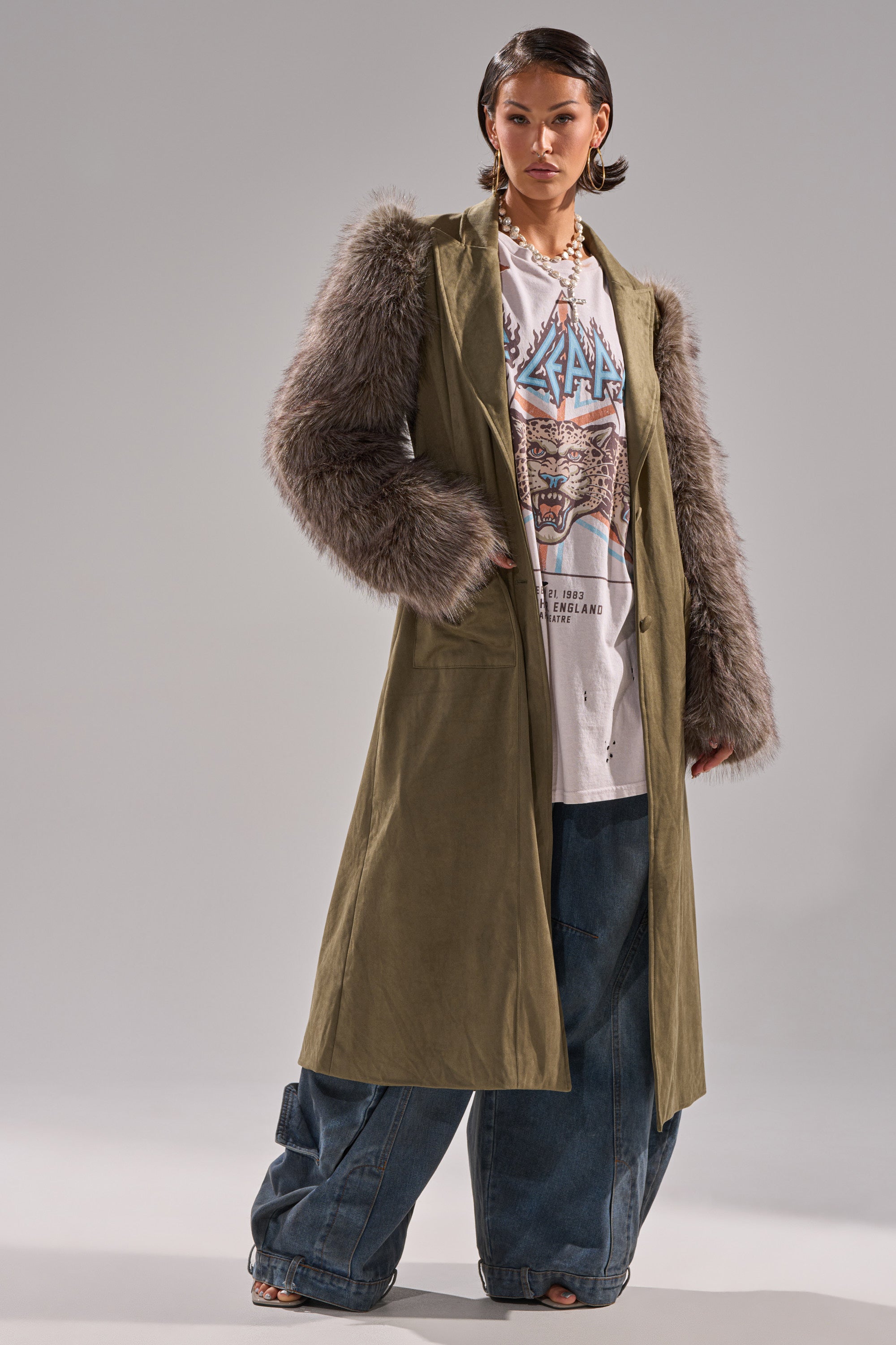 A person models the LOLITA FUR SLEEVE TRENCH with an oversized graphic tee and wide-leg blue jeans, achieving a trendy and eclectic look against a plain backdrop.