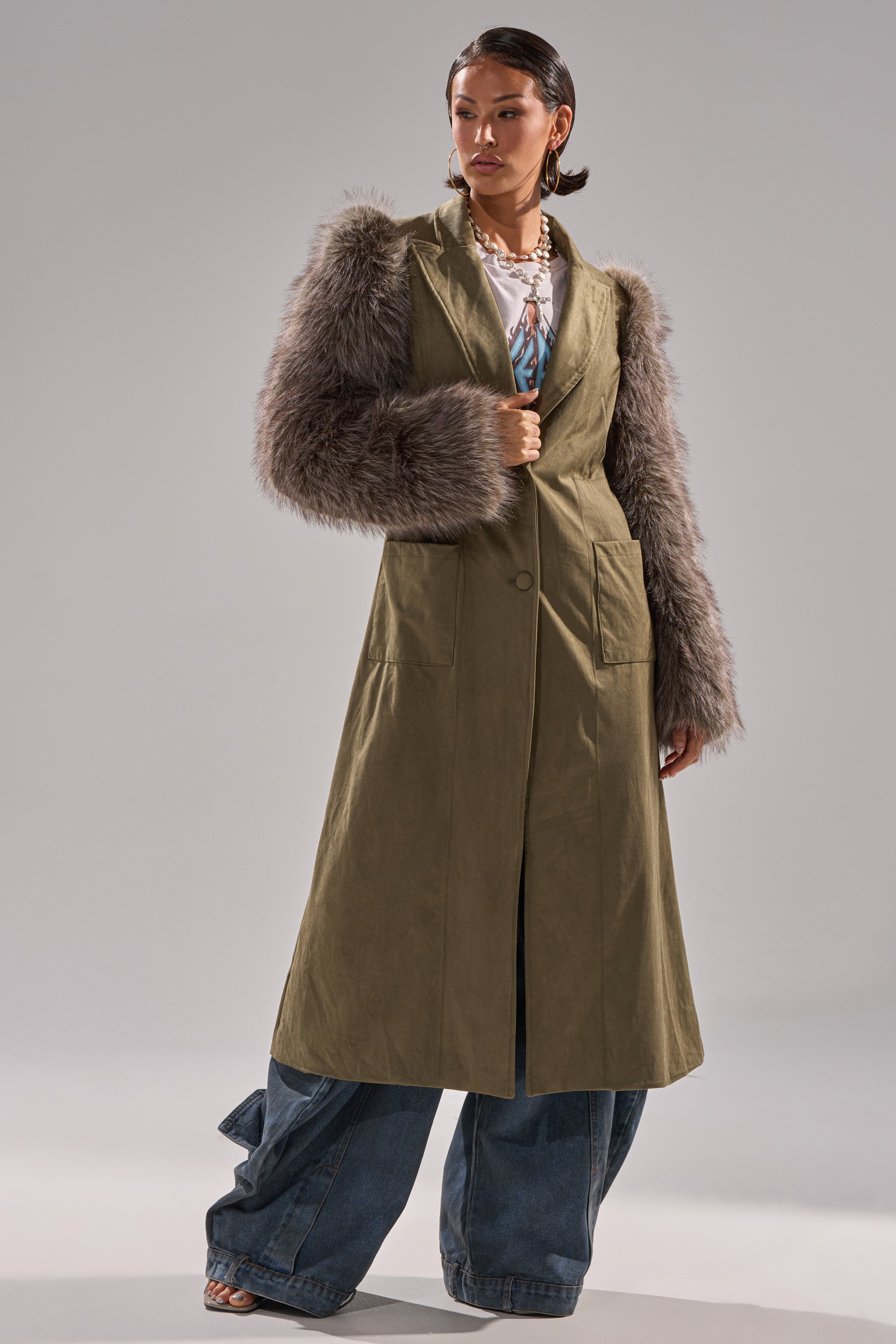 A woman models the LOLITA FUR SLEEVE TRENCH, an olive midi coat with shaggy gray fur sleeves, paired with wide-leg blue jeans and layered necklaces, against a plain gray background.