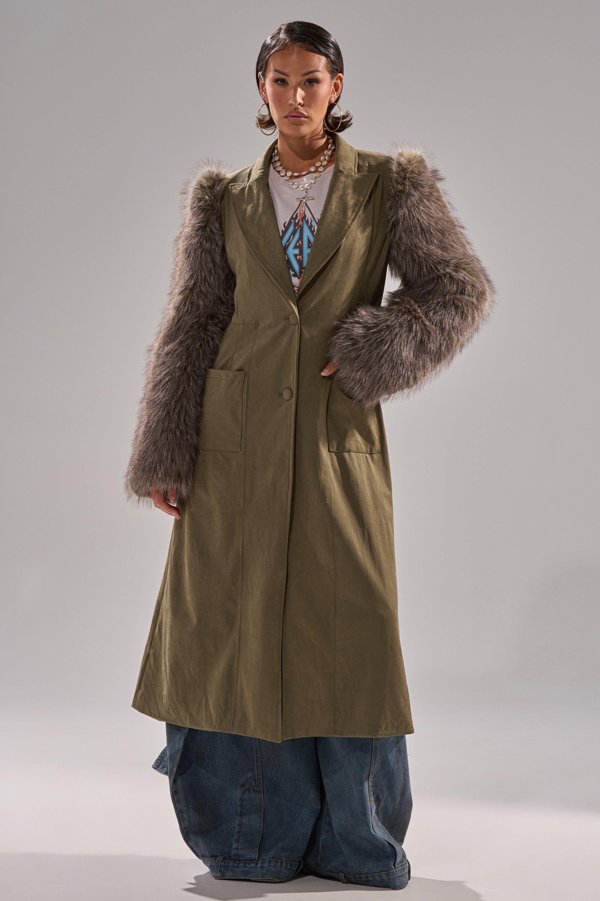 A person wears the LOLITA FUR SLEEVE TRENCH in olive green with faux fur sleeves, styled with a graphic tee, layered necklaces, and wide-leg blue jeans, standing against a plain light background.