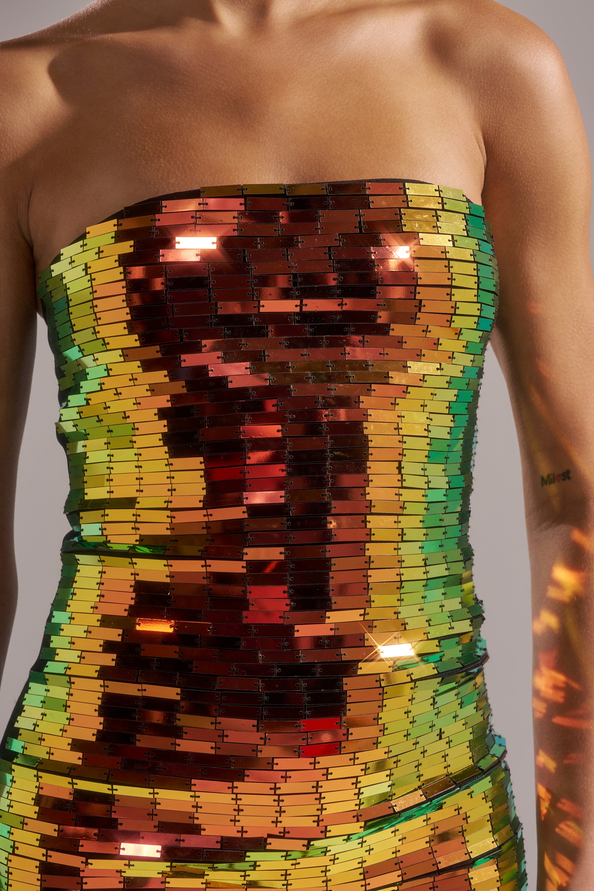 Wearing the DANCIN THE NIGHT AWAY MINI DRESS, a strapless sequin mini reflecting gold, green, and reddish-orange shades. The upper chest and arms are visible, with a small right arm tattoo.