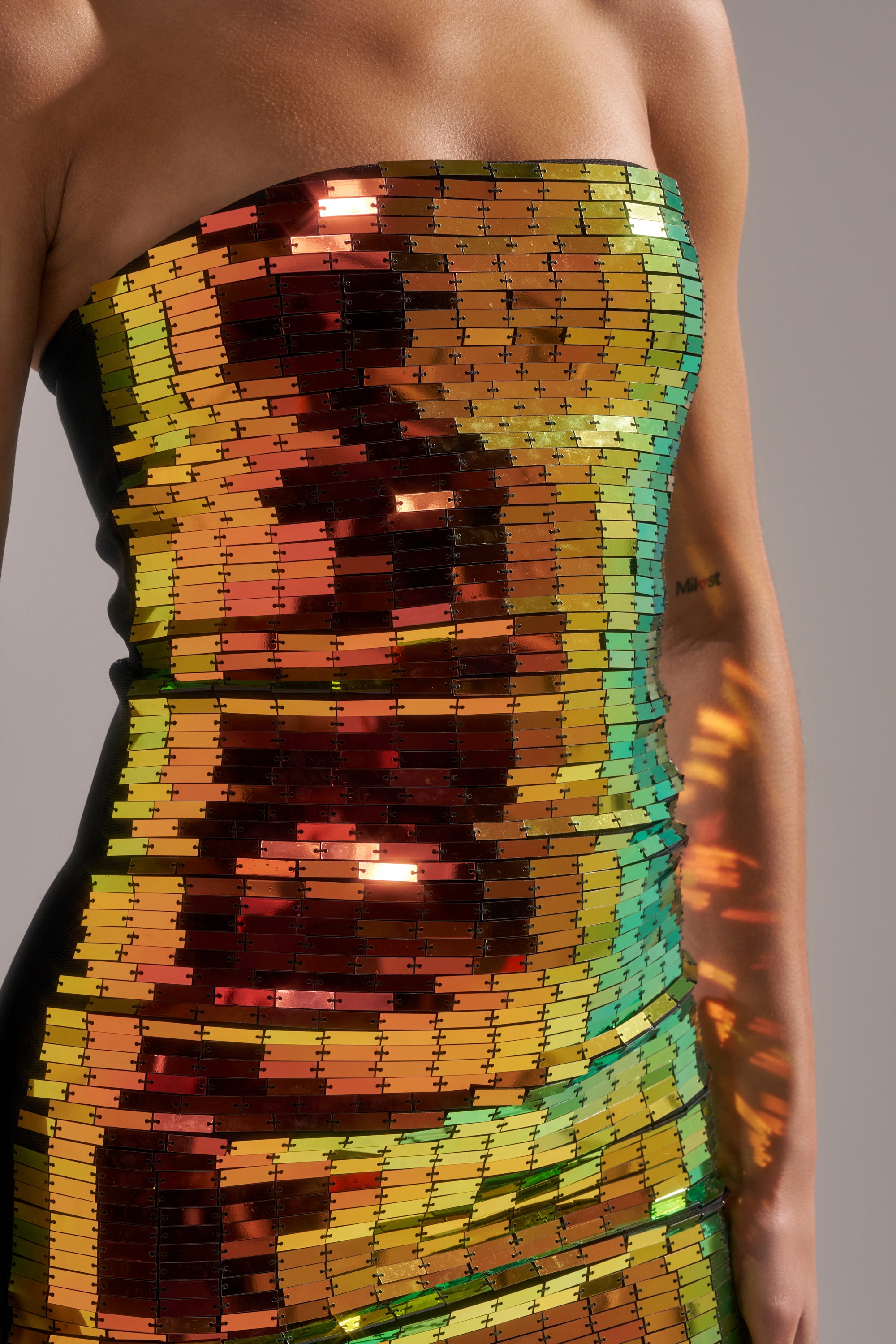 A person wearing the DANCIN THE NIGHT AWAY MINI DRESS, a strapless dress adorned with shiny, rectangular sequins in red, orange, yellow, and green hues. Only the torso and part of one arm are visible against a neutral background.