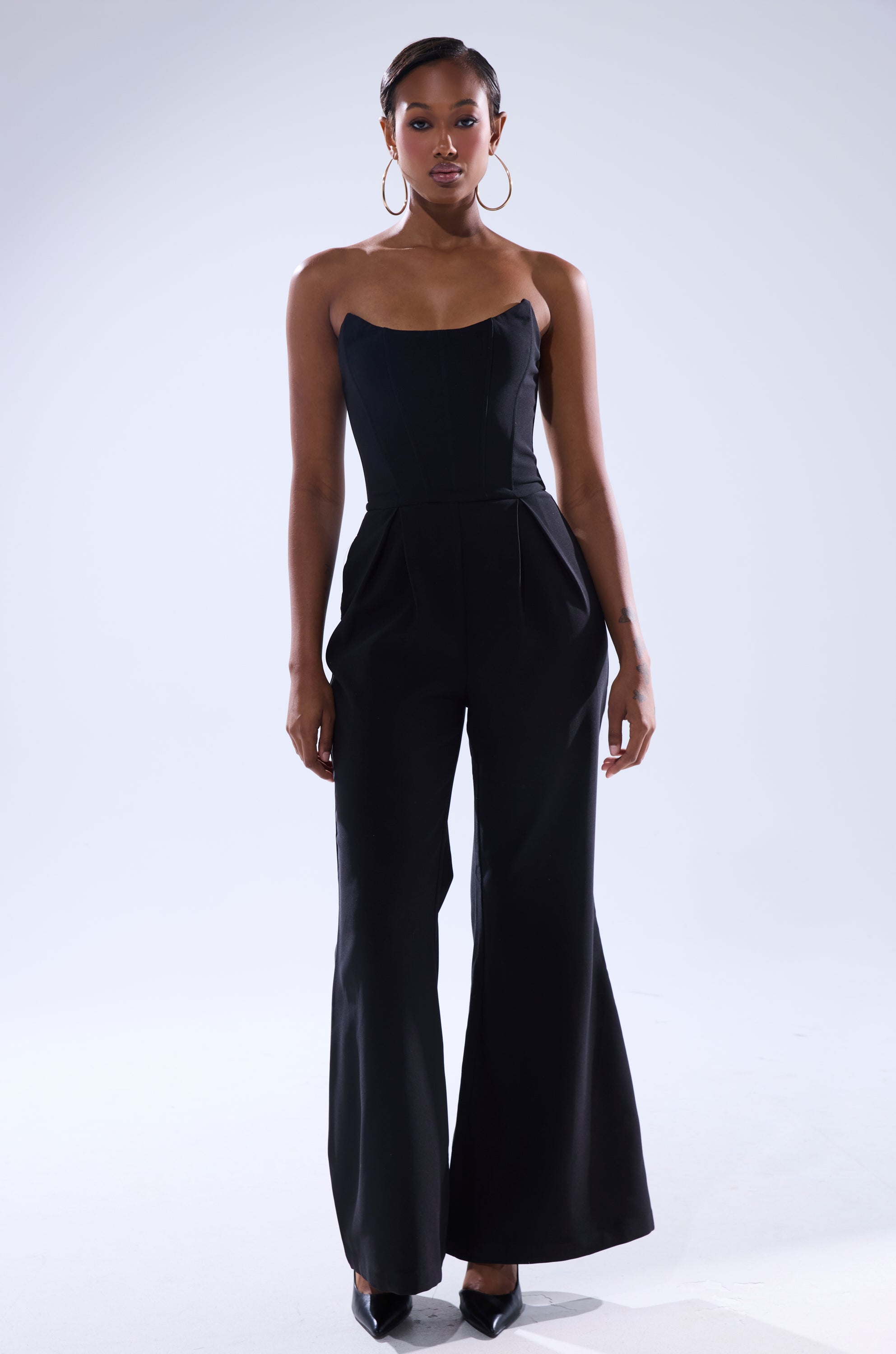 A woman stands against a plain light background, wearing the LOVELY DAYS JUMPSUIT IN BLACK—a strapless, wide-leg jumpsuit paired with black heels, hoop earrings, and her hair styled back.