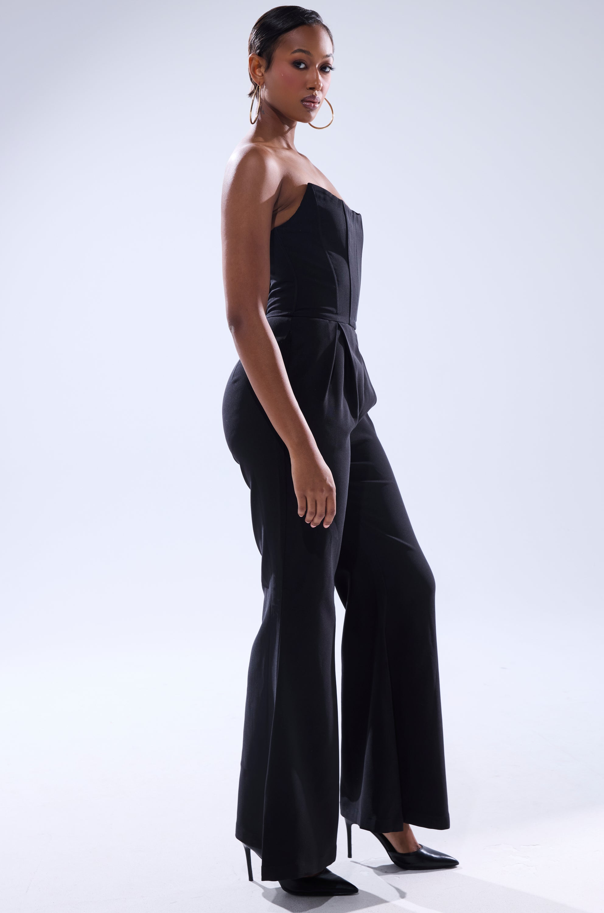 A woman poses confidently in the LOVELY DAYS JUMPSUIT IN BLACK from AKIRA Label, paired with black high heels and hoop earrings. Her short, sleek hair and subtle body turn stand out against the light background.