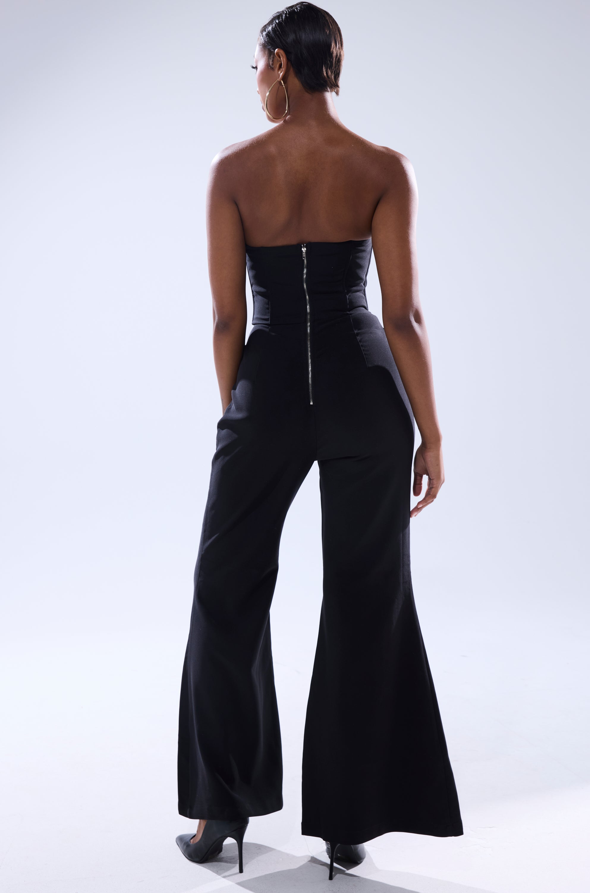A woman faces away, wearing the LOVELY DAYS JUMPSUIT IN BLACK by AKIRA Label—strapless, with wide-leg pants and a visible back zipper—styled with short hair, large hoop earrings, and black heels against a light background.
