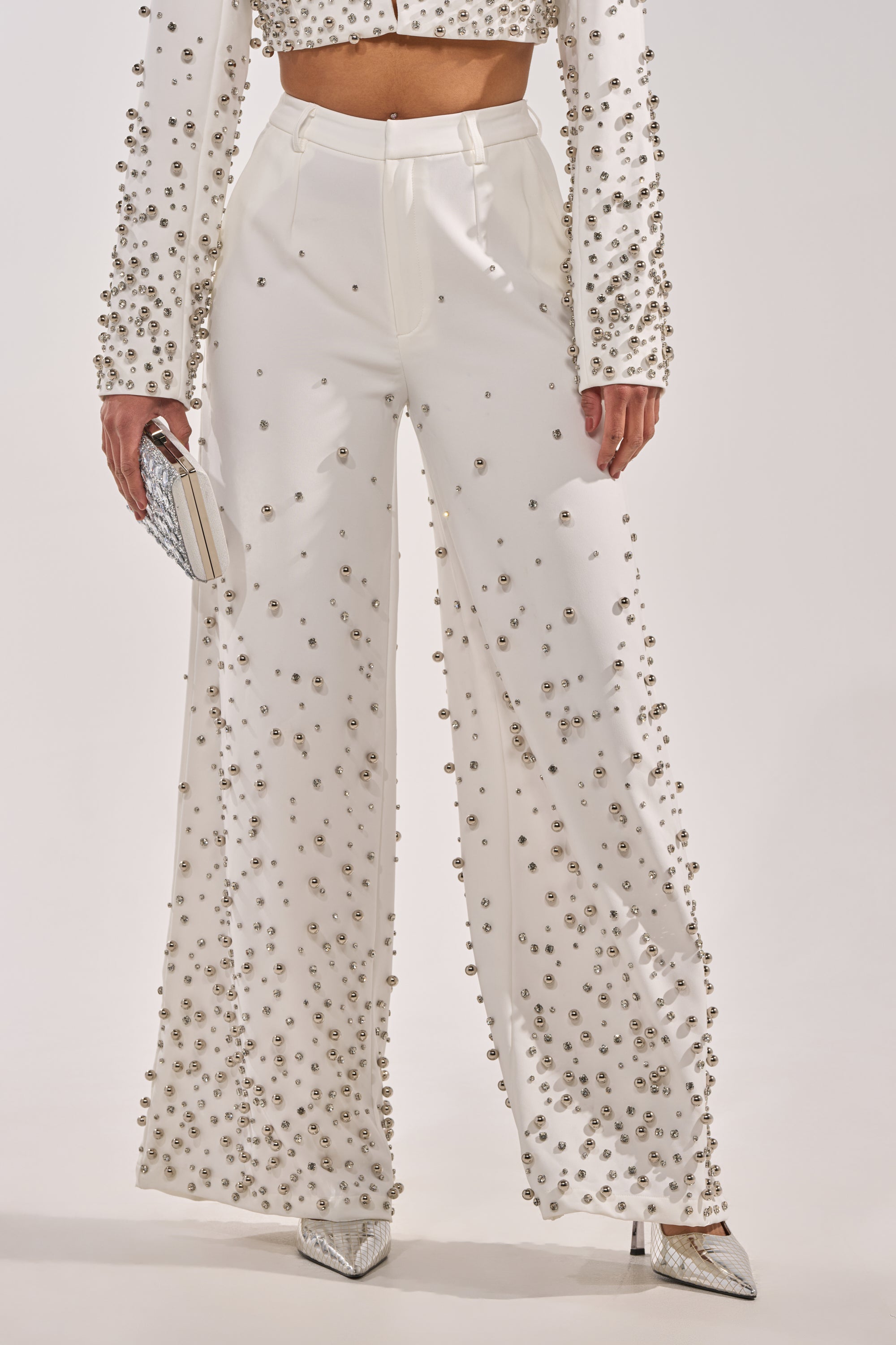 Wearing the GET CLOSER EMBELLISHED TROUSER IN WHITE, styled with a matching silver-embellished top, a small silver clutch, and pointed silver shoes. 1
