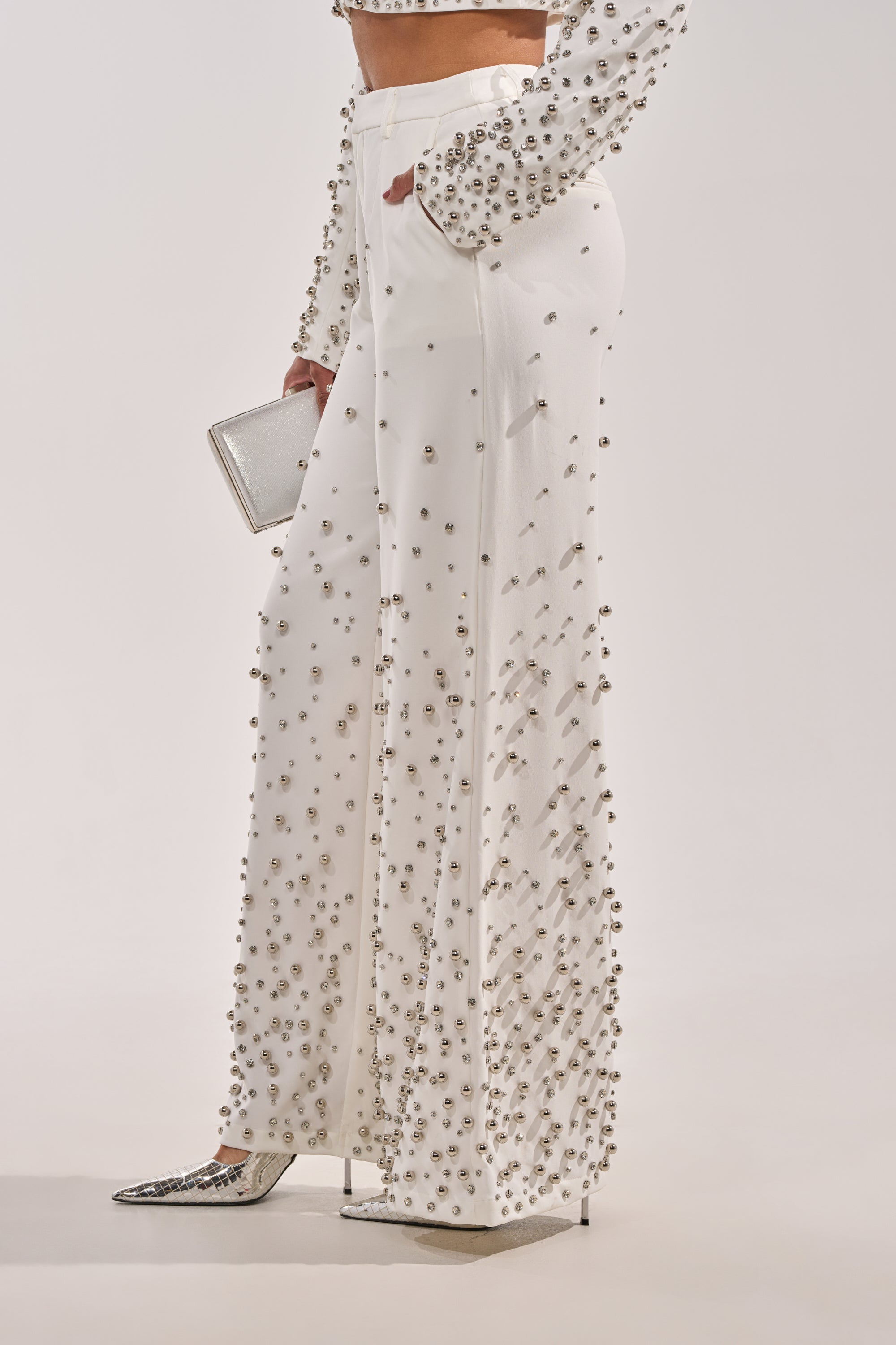 Wearing the GET CLOSER EMBELLISHED TROUSER IN WHITE with a beaded crop top, silver pointed heels, and a small silver clutch against a plain background. 2
