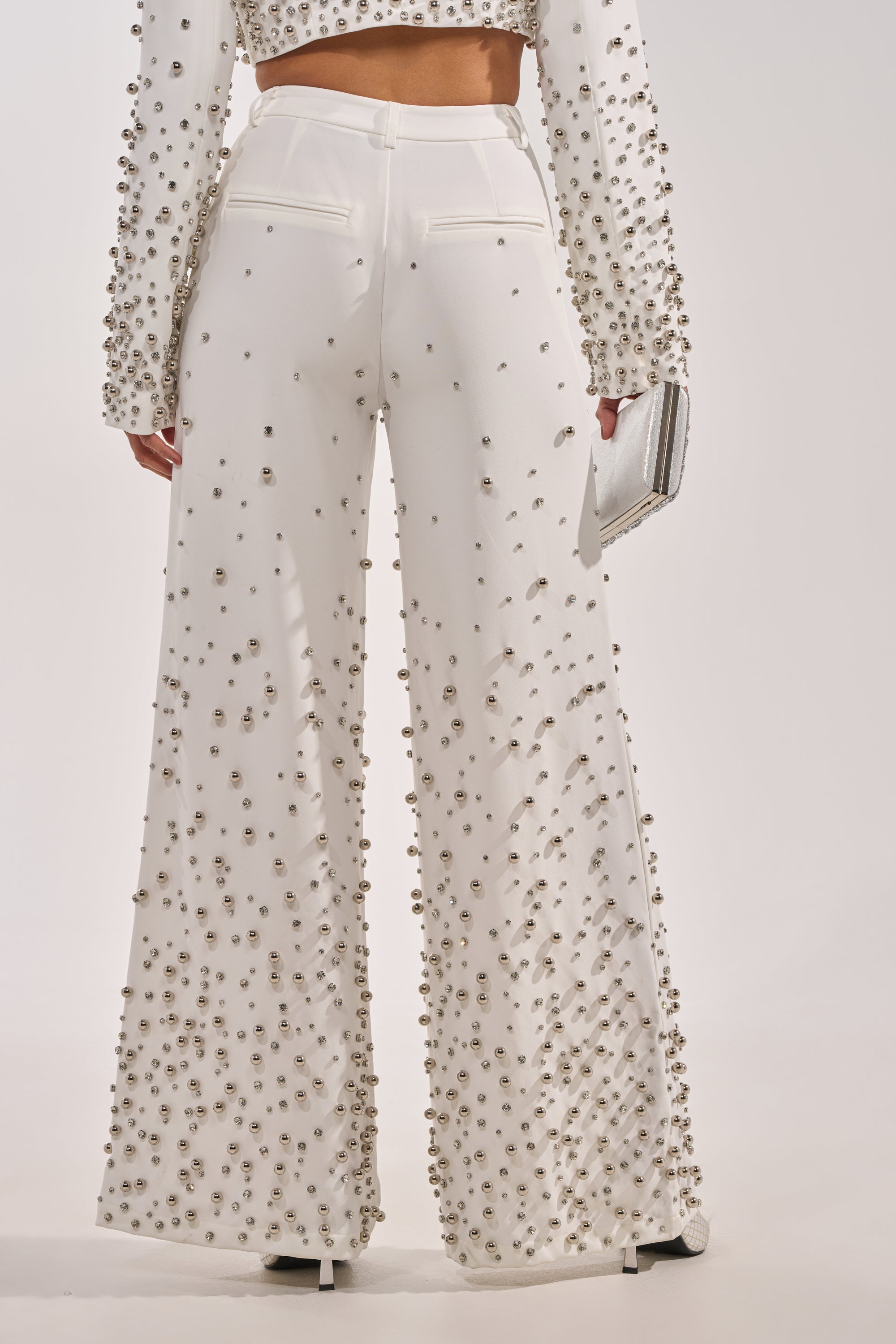 A person is seen from the back wearing the GET CLOSER EMBELLISHED TROUSER IN WHITE—high-waisted, wide-leg pants with rhinestone detailing—and holding a silver clutch, showcasing the intricate embellishments. 3