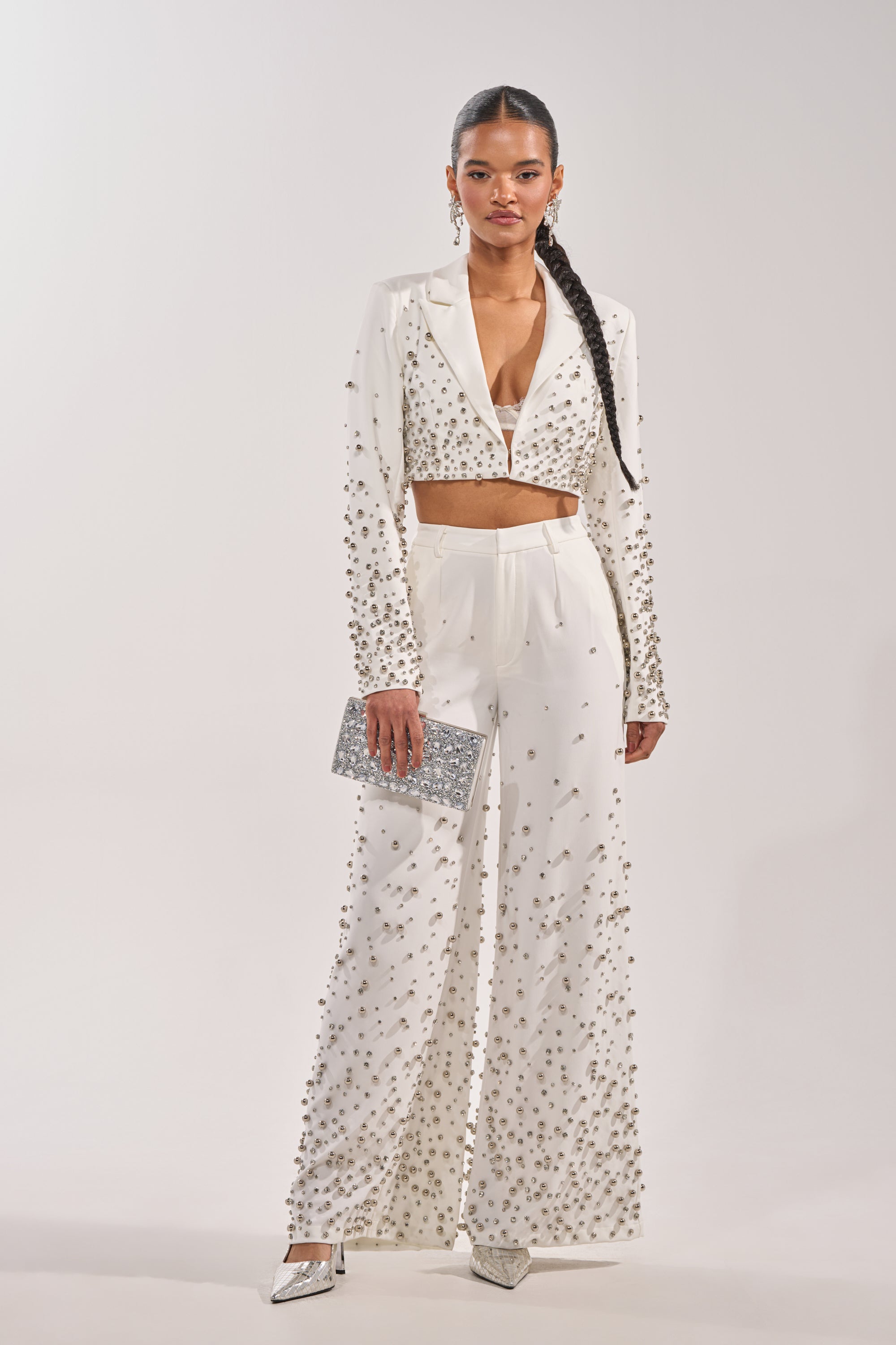 A woman wears the GET CLOSER EMBELLISHED TROUSER IN WHITE with a matching cropped blazer, accessorized with a silver clutch and pointed silver heels. Her hair is styled in a long braid. 4