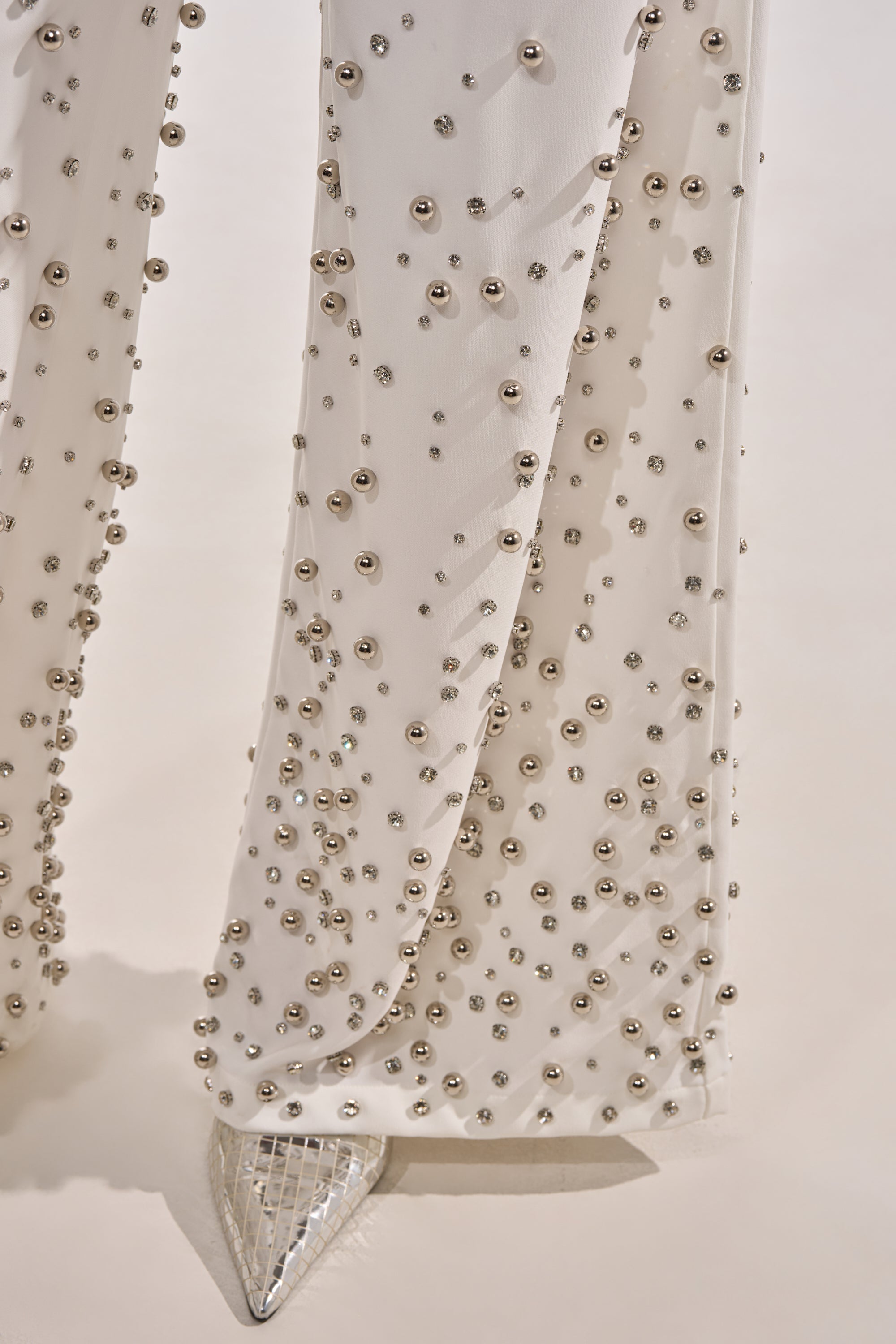 The GET CLOSER EMBELLISHED TROUSER IN WHITE, adorned with metallic beads, is styled with pointed silver geometric shoes. The photo highlights the lower legs against a light background. 5