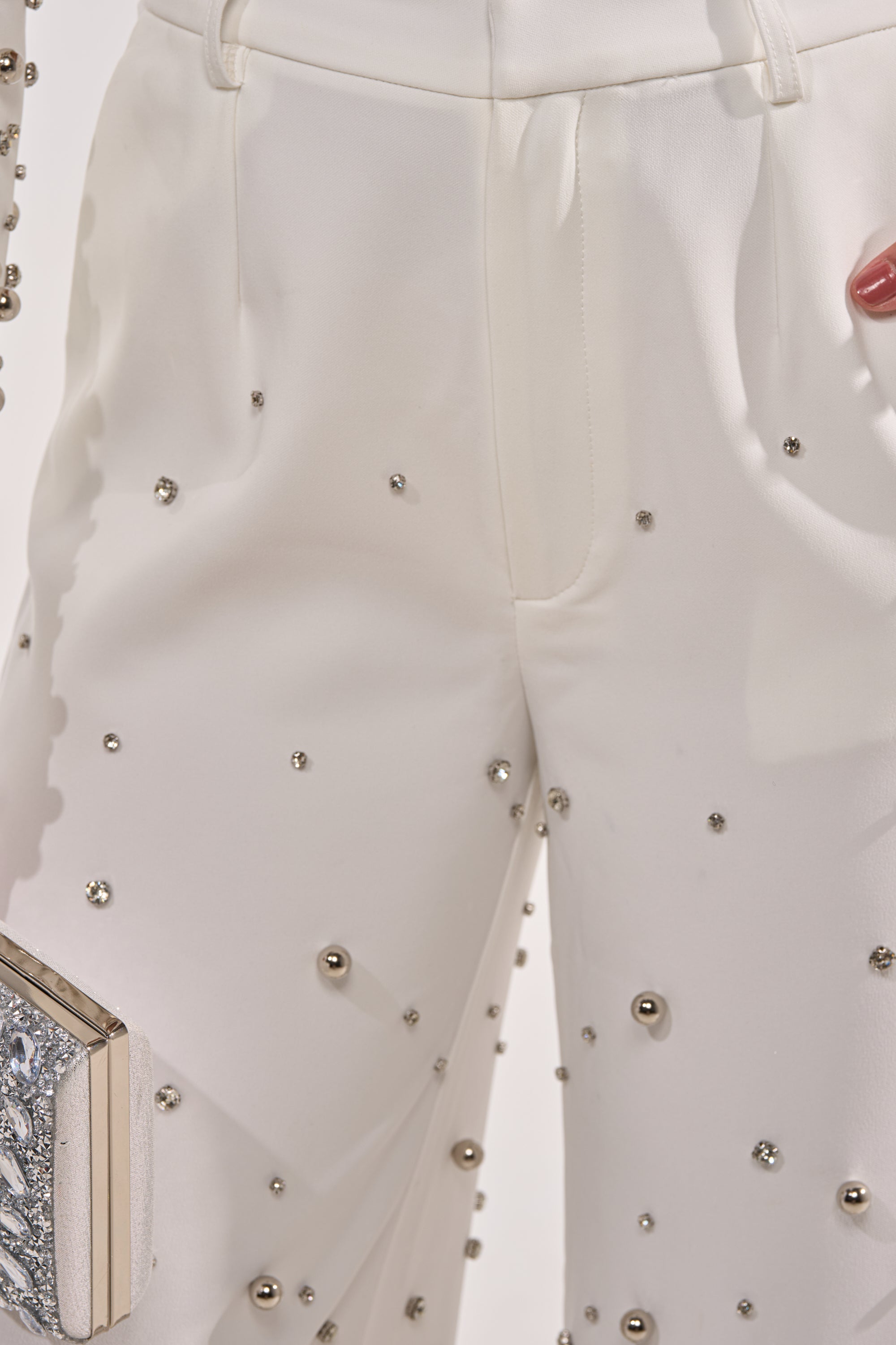 A close-up of a person wearing the GET CLOSER EMBELLISHED TROUSER IN WHITE, detailed with metallic studs. They hold a textured silver clutch, and their nails are painted red. 6