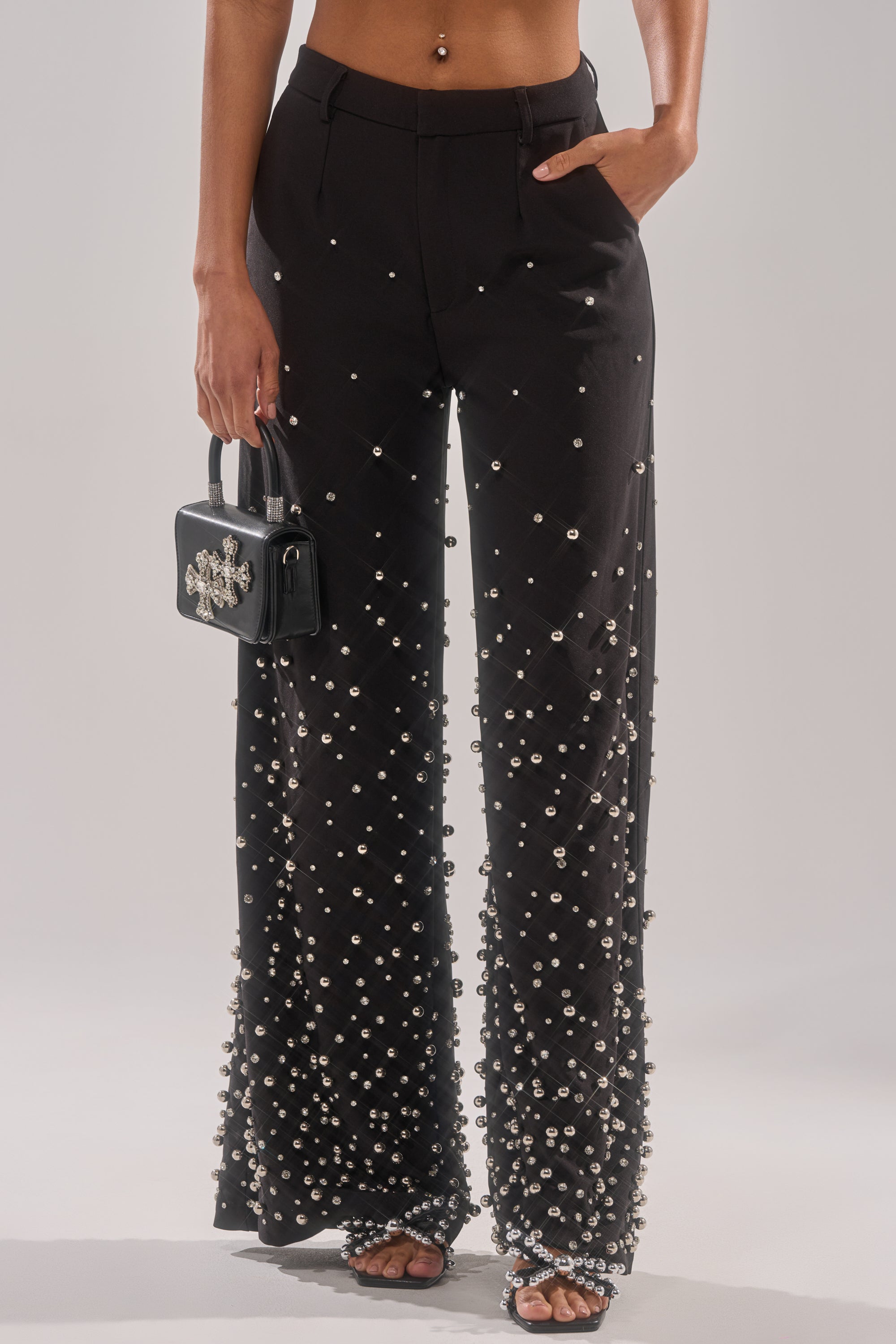 Wearing the GET CLOSER EMBELLISHED TROUSER—high-waisted, wide-leg black pants with silver pearls—paired with a small black handbag featuring a silver design and open-toe heeled sandals. Their midriff and belly piercing are visible.