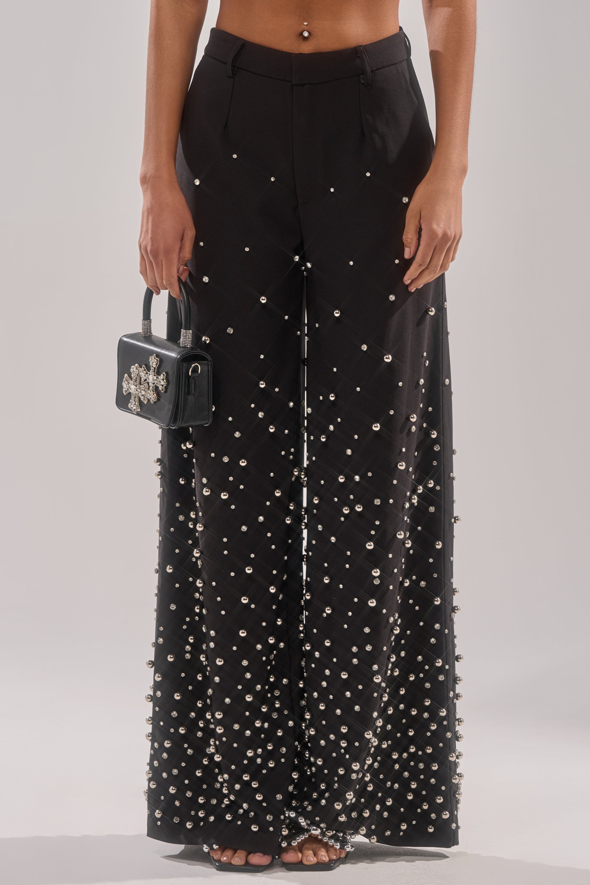 A person models the GET CLOSER EMBELLISHED TROUSER—black wide-leg pants with pearl-like beads—holding a small black embellished handbag, barefoot against a light backdrop.