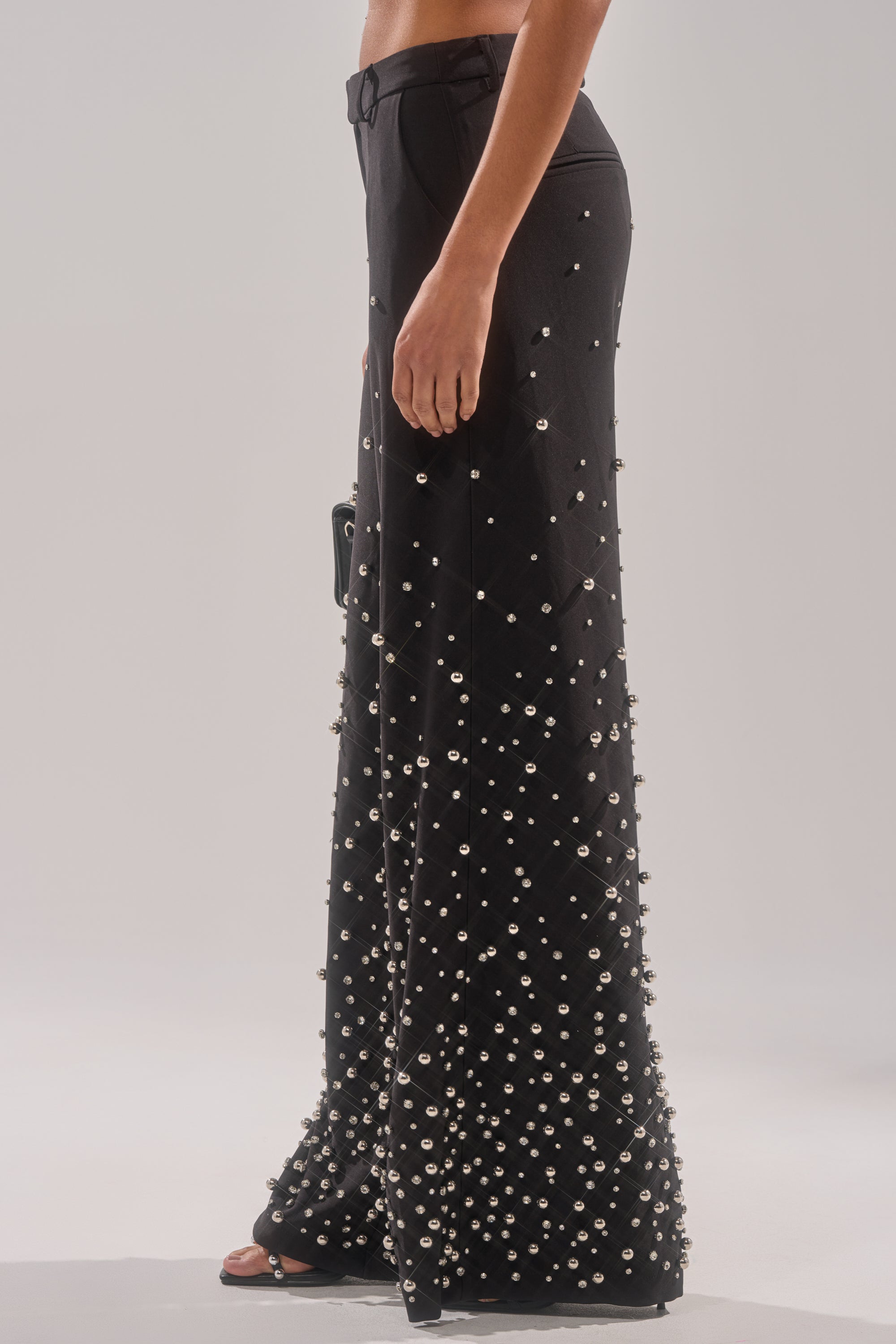 A person models the GET CLOSER EMBELLISHED TROUSER—high-waisted black pants with metallic studs concentrated near the hem—paired with open-toe black heels.
