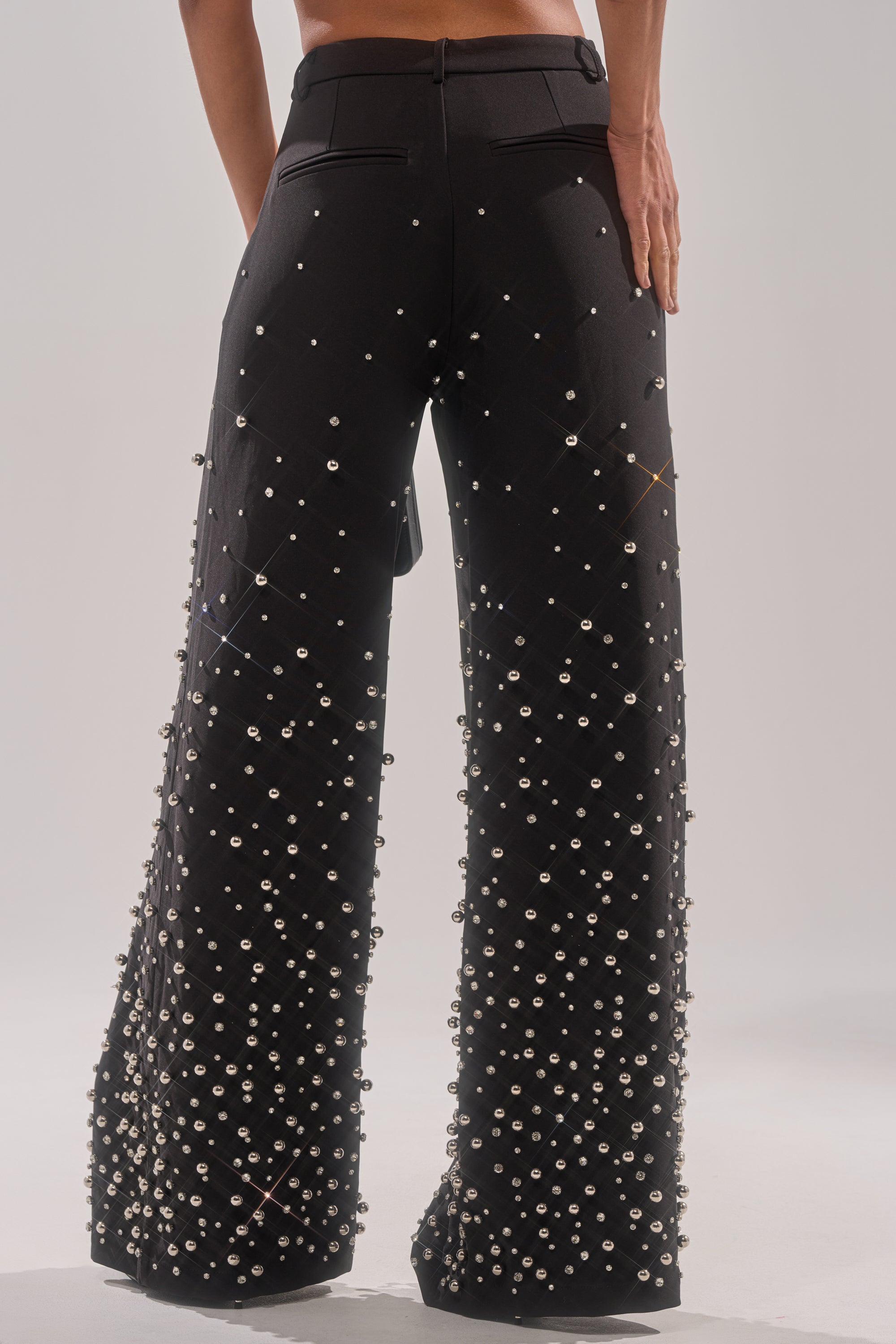 A person stands facing away, wearing the GET CLOSER EMBELLISHED TROUSER—black, high-waisted wide leg pants decorated with shiny silver studs and pearls for a striking textured look.