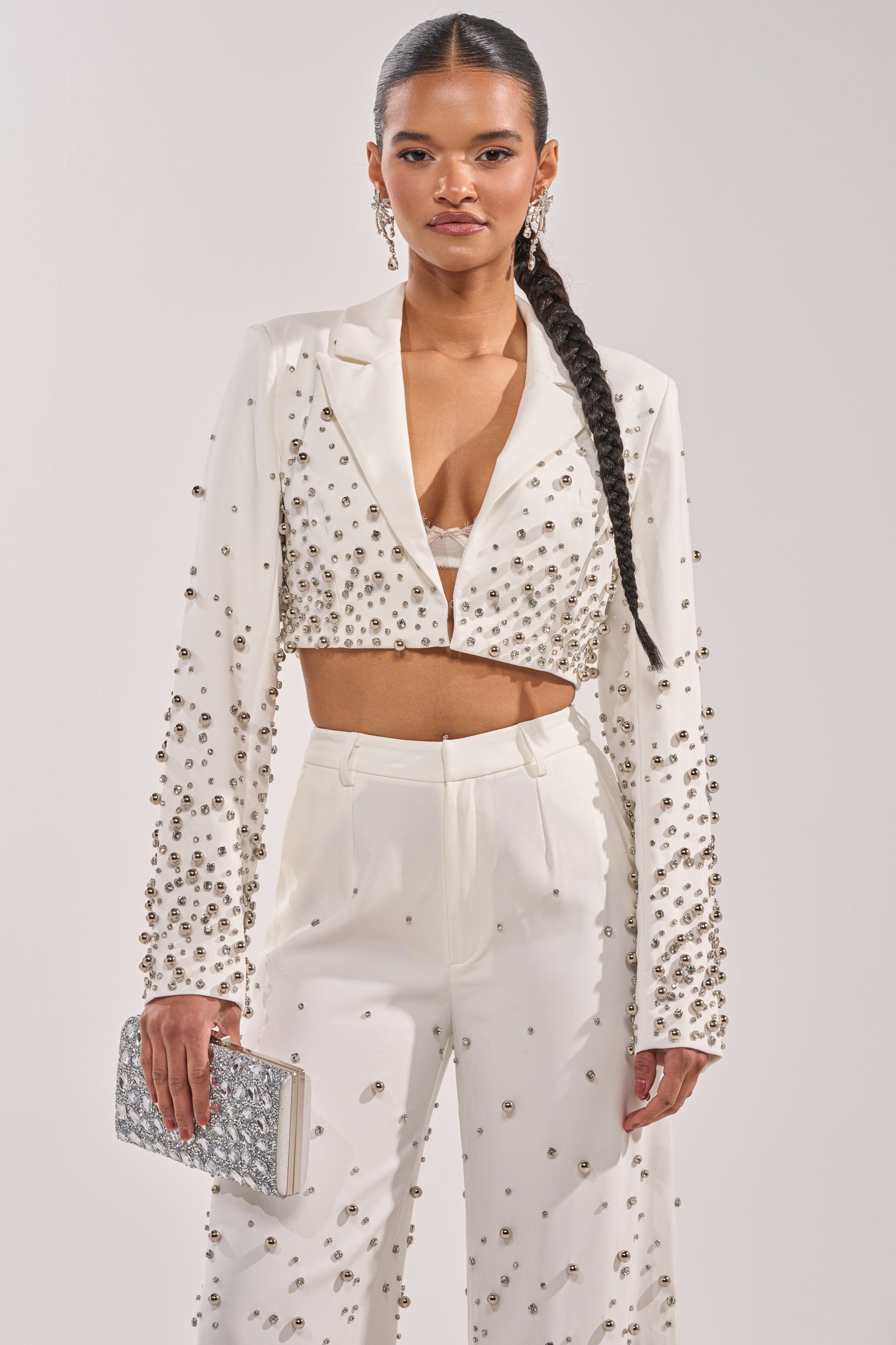 A woman with long, braided hair poses confidently in the GET CLOSER EMBELLISHED BLAZER IN WHITE and matching pants, accessorized with a silver embellished clutch and statement earrings against a plain background. 1