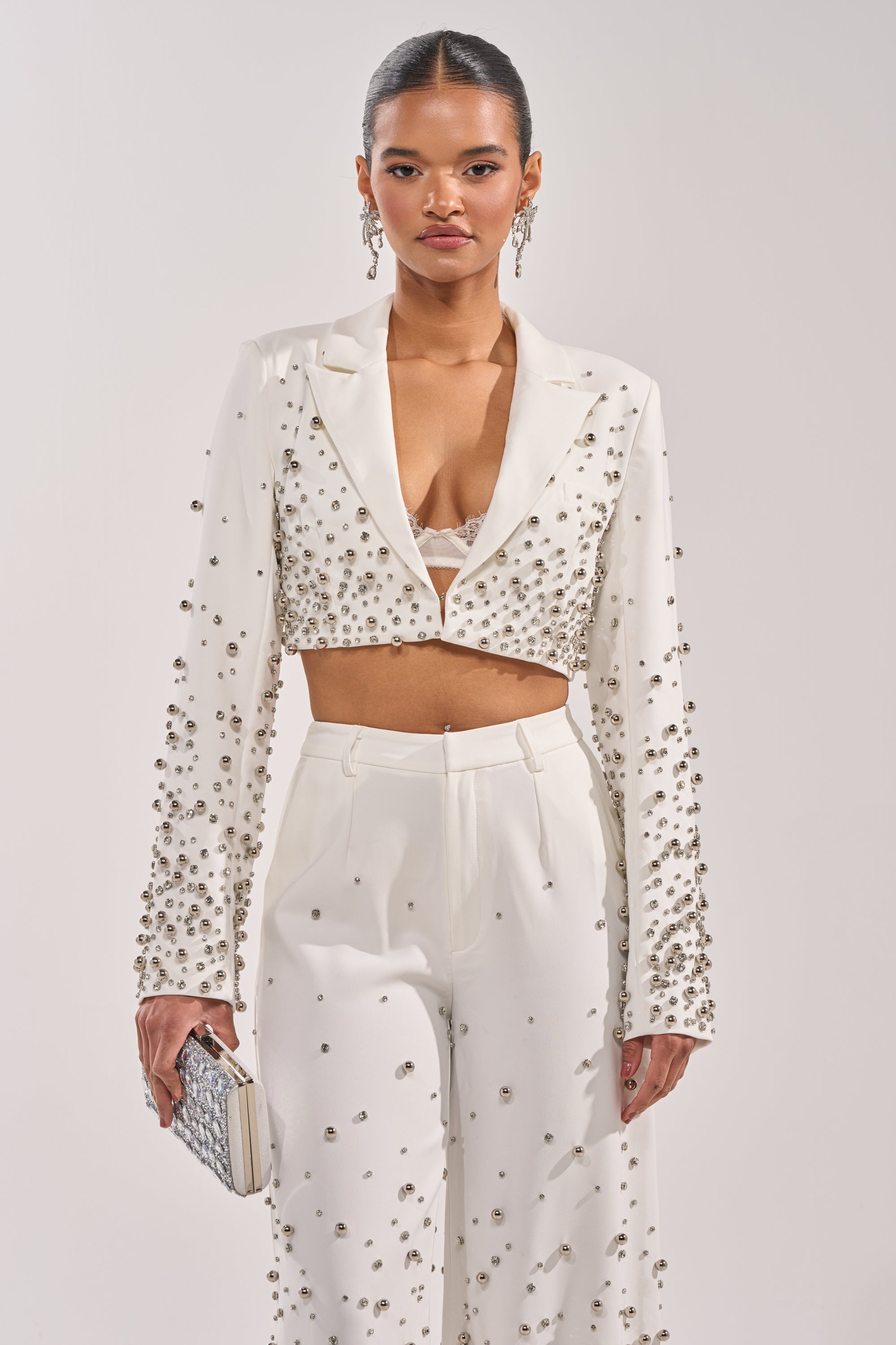 A woman confidently poses against a plain backdrop in the GET CLOSER EMBELLISHED BLAZER IN WHITE with matching high-waisted pants, accessorized with statement earrings, a sleek hairstyle, and a chic clutch. 2