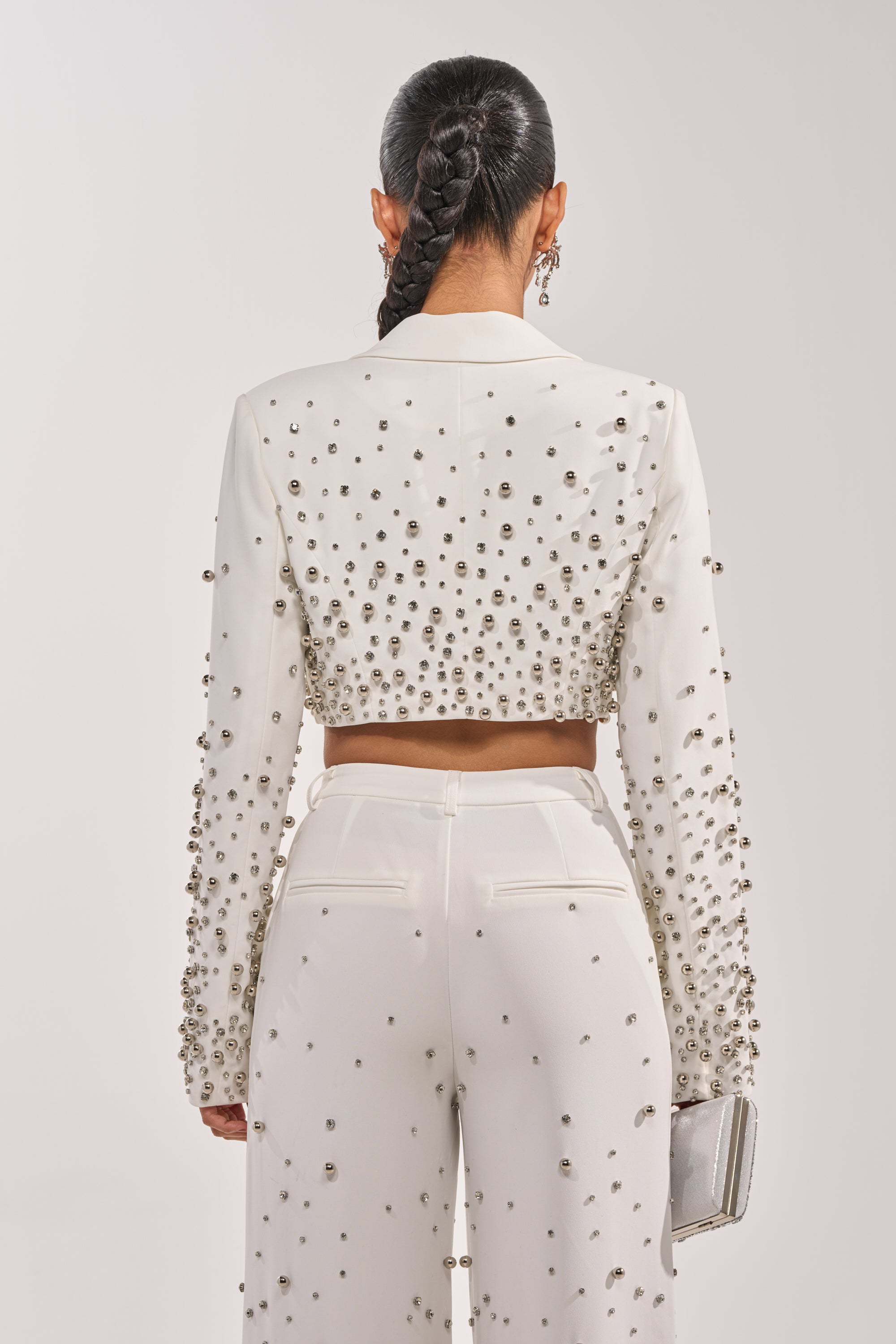 A woman with a long braided ponytail is seen from behind wearing the GET CLOSER EMBELLISHED BLAZER IN WHITE and high-waisted studded pants, holding a silver clutch. 4