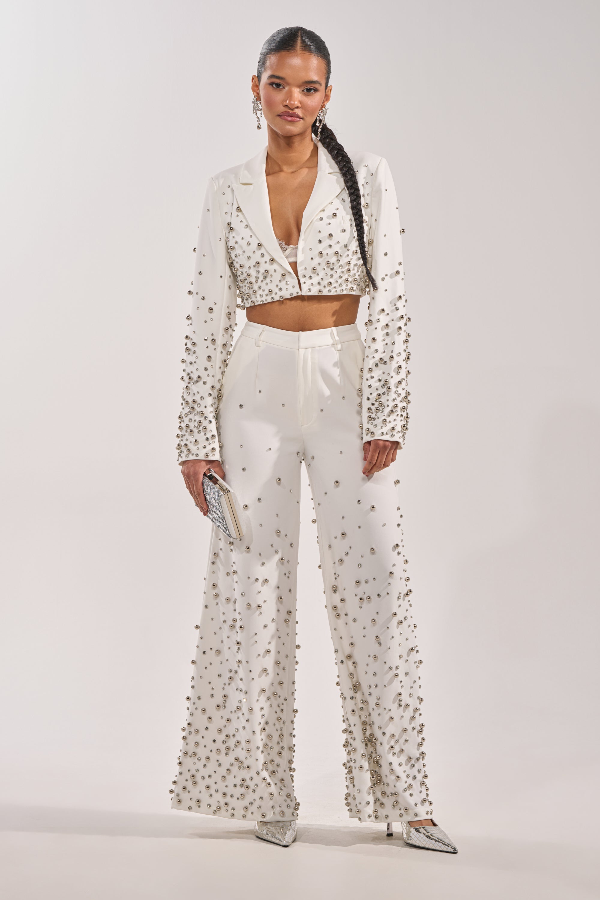 A woman stands confidently in the GET CLOSER EMBELLISHED BLAZER IN WHITE with matching wide-leg pants, paired with silver pointed heels and a clutch. Her long braid and the chic outfit create a stunning party-ready look against a plain background. 5