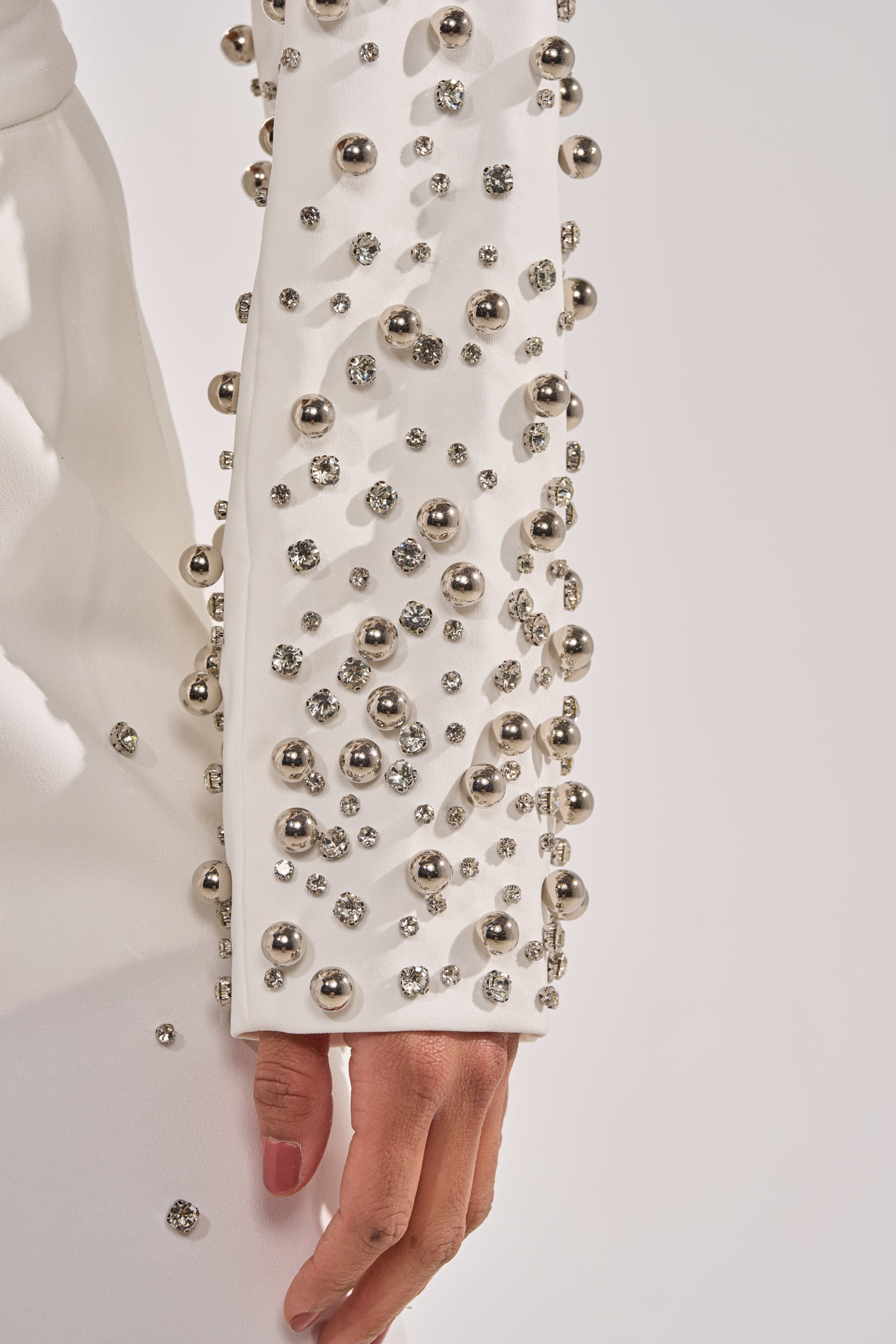 A close-up of a person’s arm wearing the GET CLOSER EMBELLISHED BLAZER IN WHITE, adorned with silver beads and rhinestones, their relaxed hand visible against a plain light background. 7