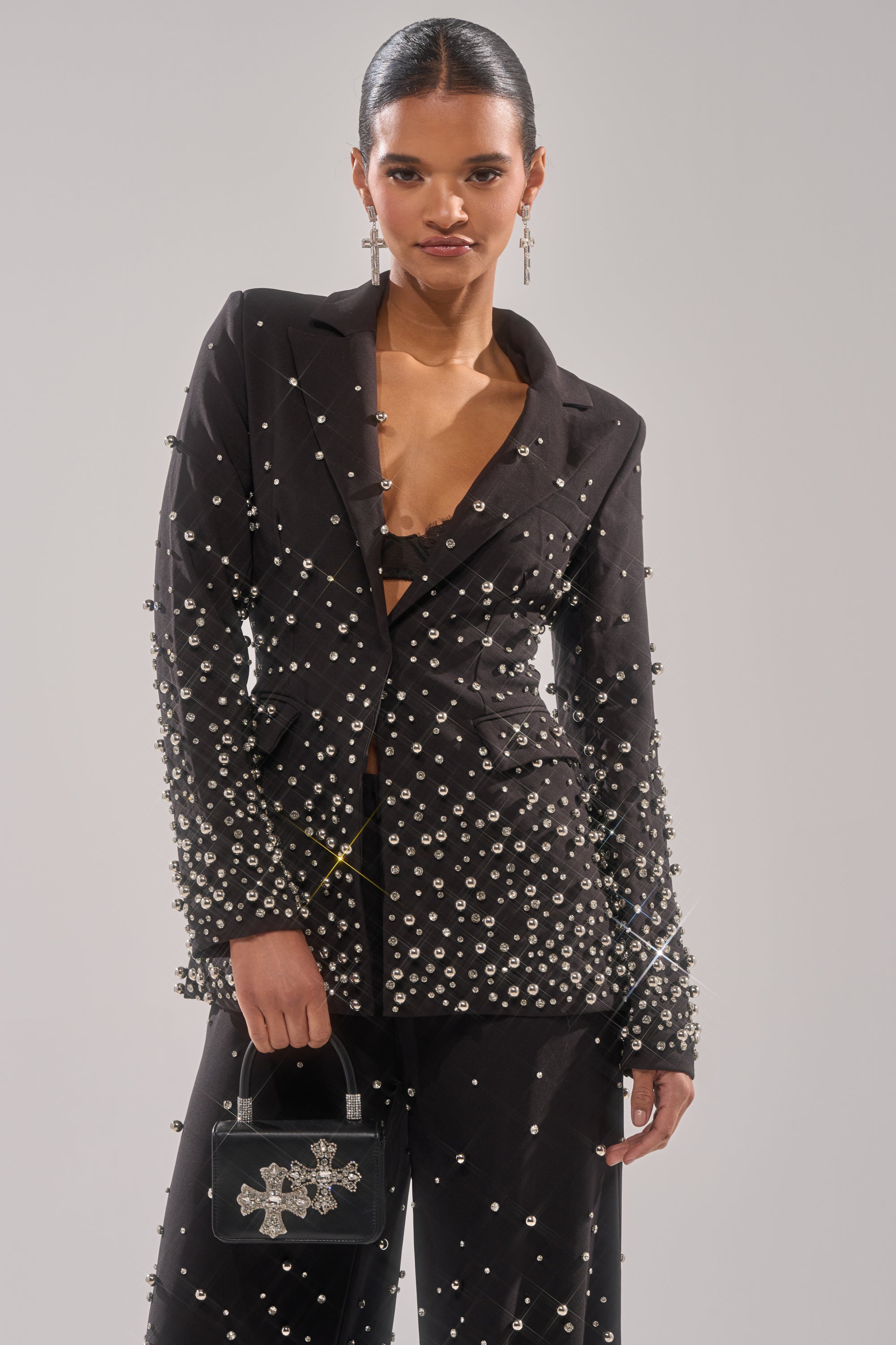 A woman poses in the fitted GET CLOSER EMBELLISHED BLAZER adorned with pearl-like beads. She holds a small black handbag with a decorative cross, wears dangling earrings, and has her hair slicked back.