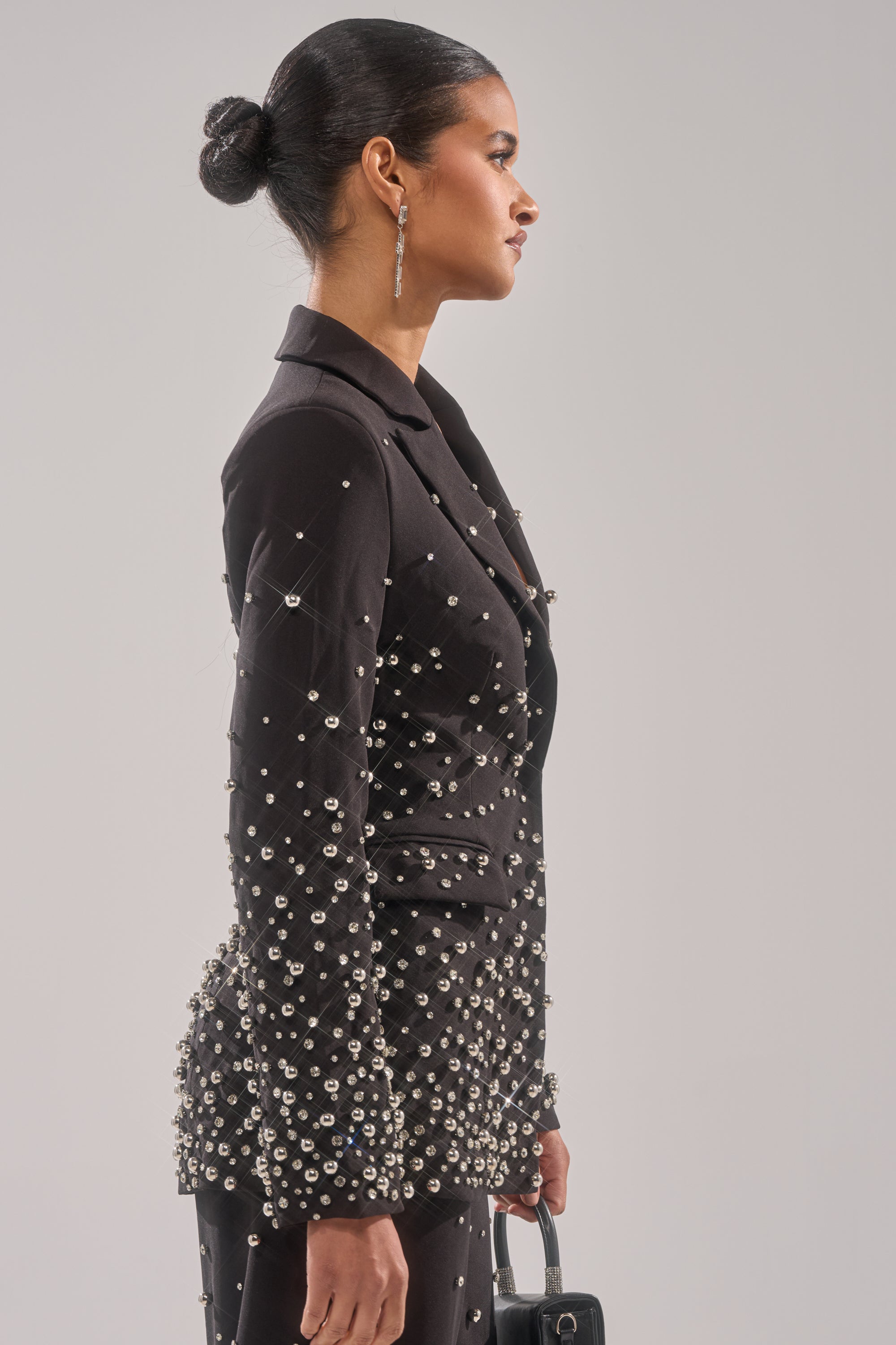 A woman with her hair in a sleek bun, wearing the GET CLOSER EMBELLISHED BLAZER, stands in profile holding a small black handbag against a plain background.