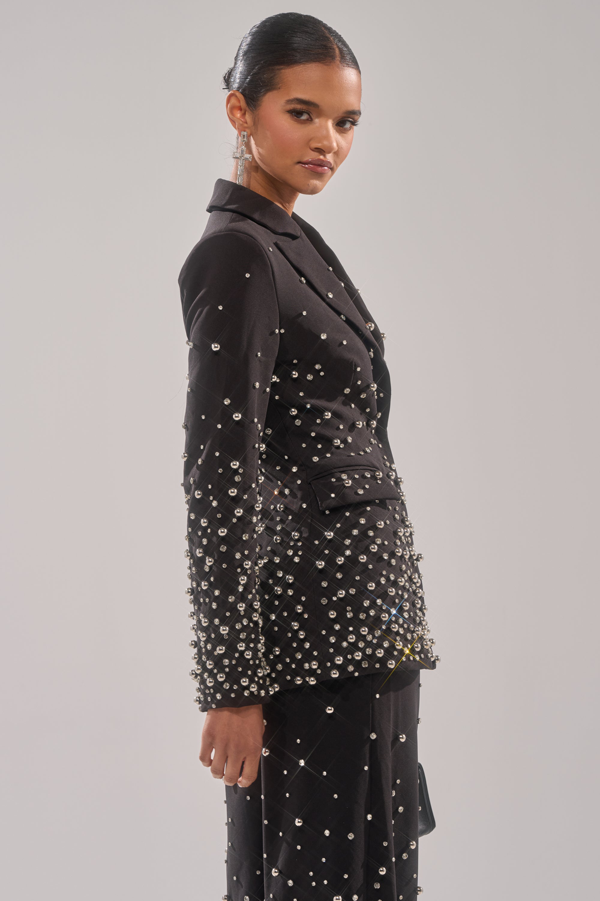 A woman poses in the GET CLOSER EMBELLISHED BLAZER, featuring sparkling silver rhinestones. She stands before a light gray background with her hair in a sleek bun and statement earrings accentuating the look.