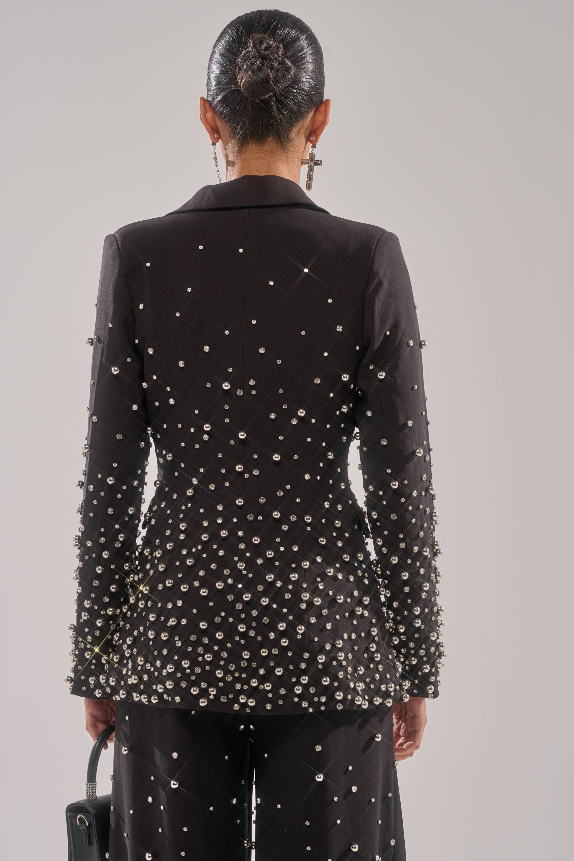 A person with dark hair in a sleek bun, seen from the back, wears the GET CLOSER EMBELLISHED BLAZER and matching studded pants while holding a black handbag against a neutral background.