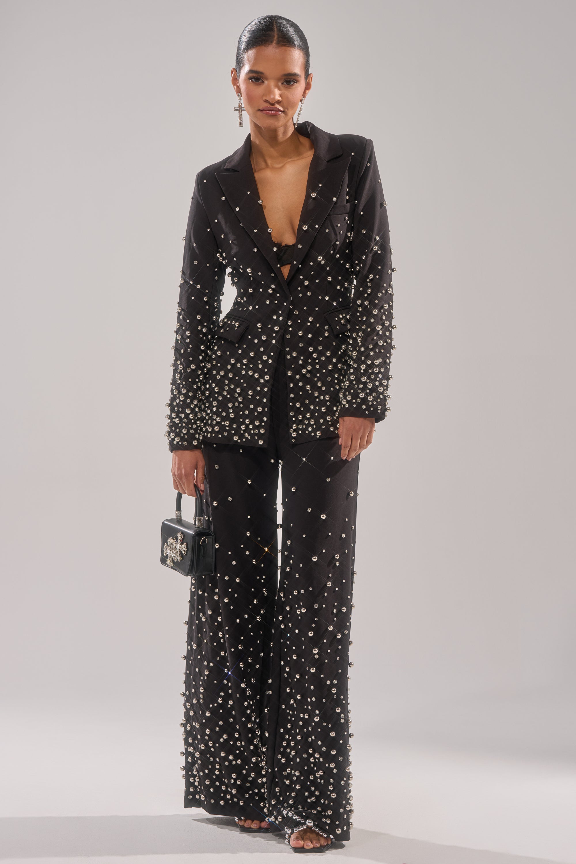 A woman stands against a plain background wearing the GET CLOSER EMBELLISHED BLAZER with black pants, a black bralette, heeled sandals, and a small black handbag. Her hair is slicked back.