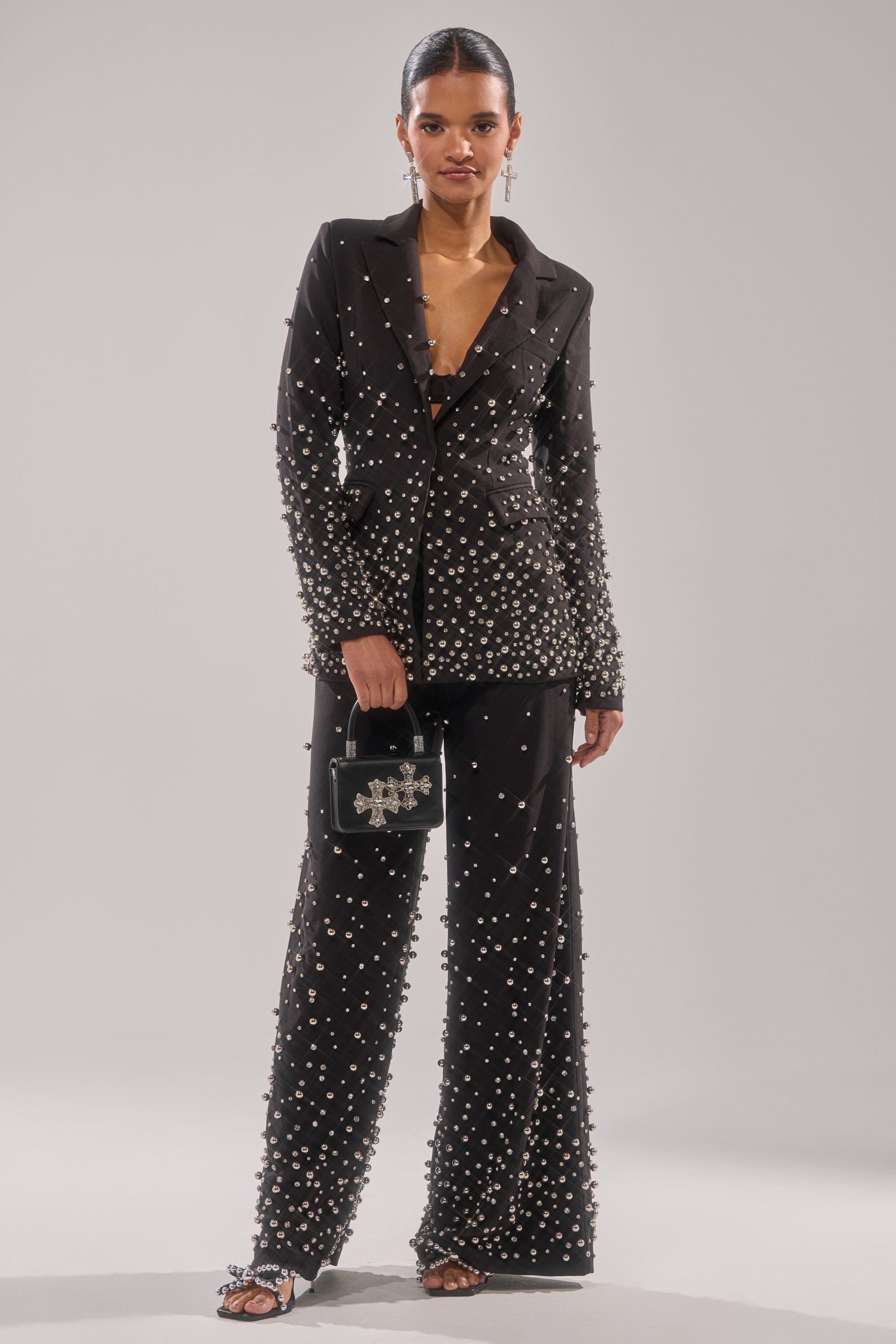 A woman stands confidently in the GET CLOSER EMBELLISHED BLAZER with matching pearl-embellished sandals, holding a small black handbag with a metallic cross. Her hair is slicked back and she wears dangling earrings.